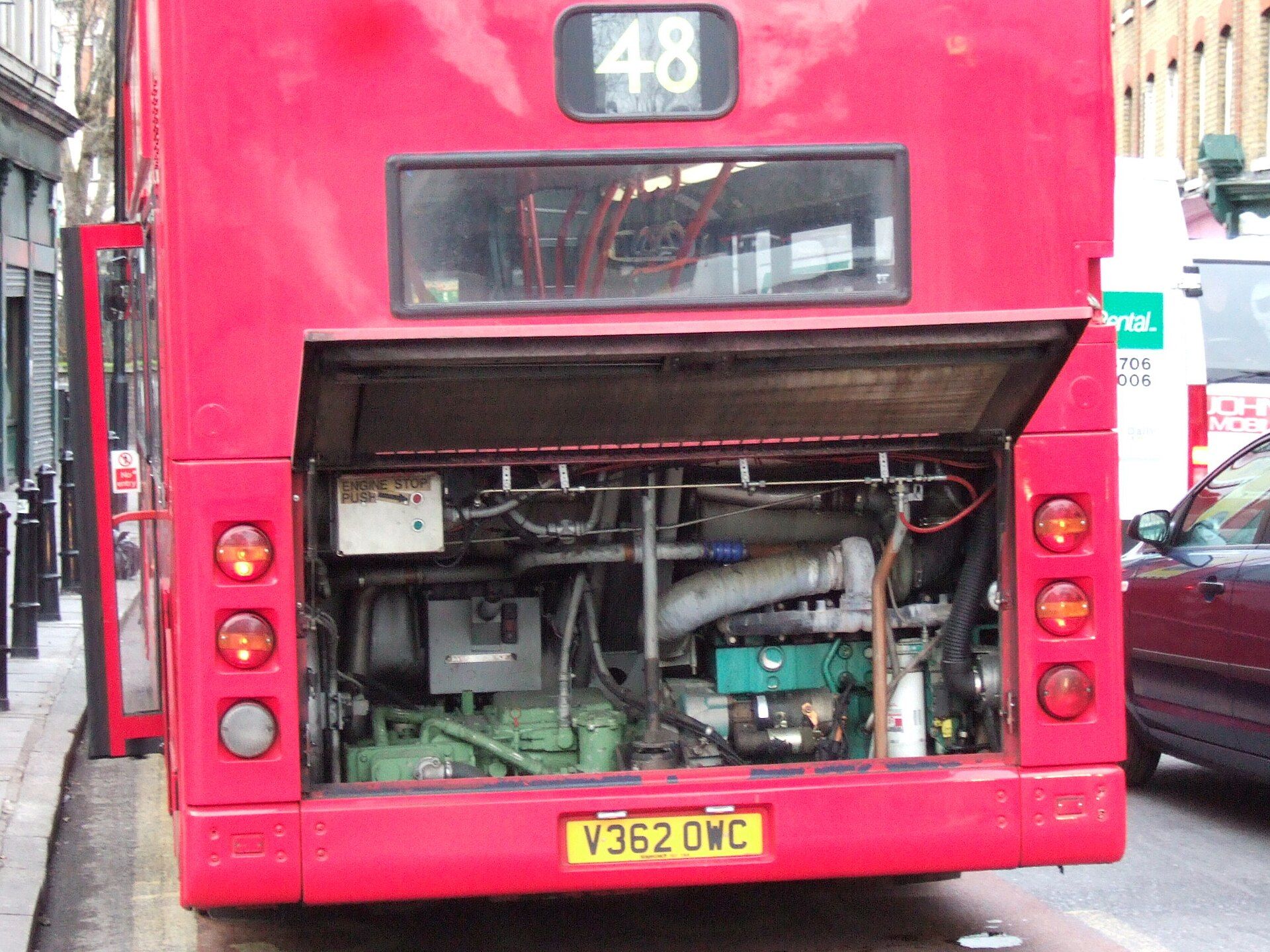 Rear engine compartment flap raised on a London double-deck bus, exposing the engine, exhaust pipework and an engine stop panel
