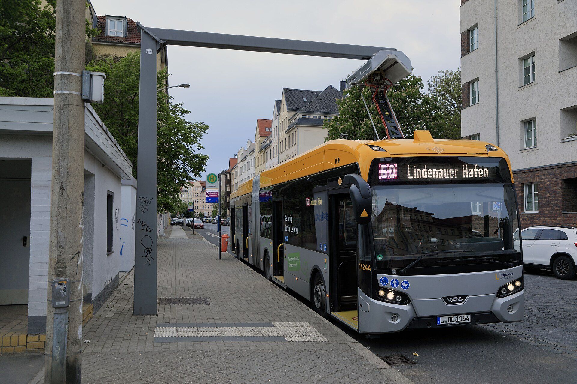 Yellow and grey articulated electric city bus at a bus stop in Leipzig, with a pantograph arm reaching from its roof up to an overhead charging mast