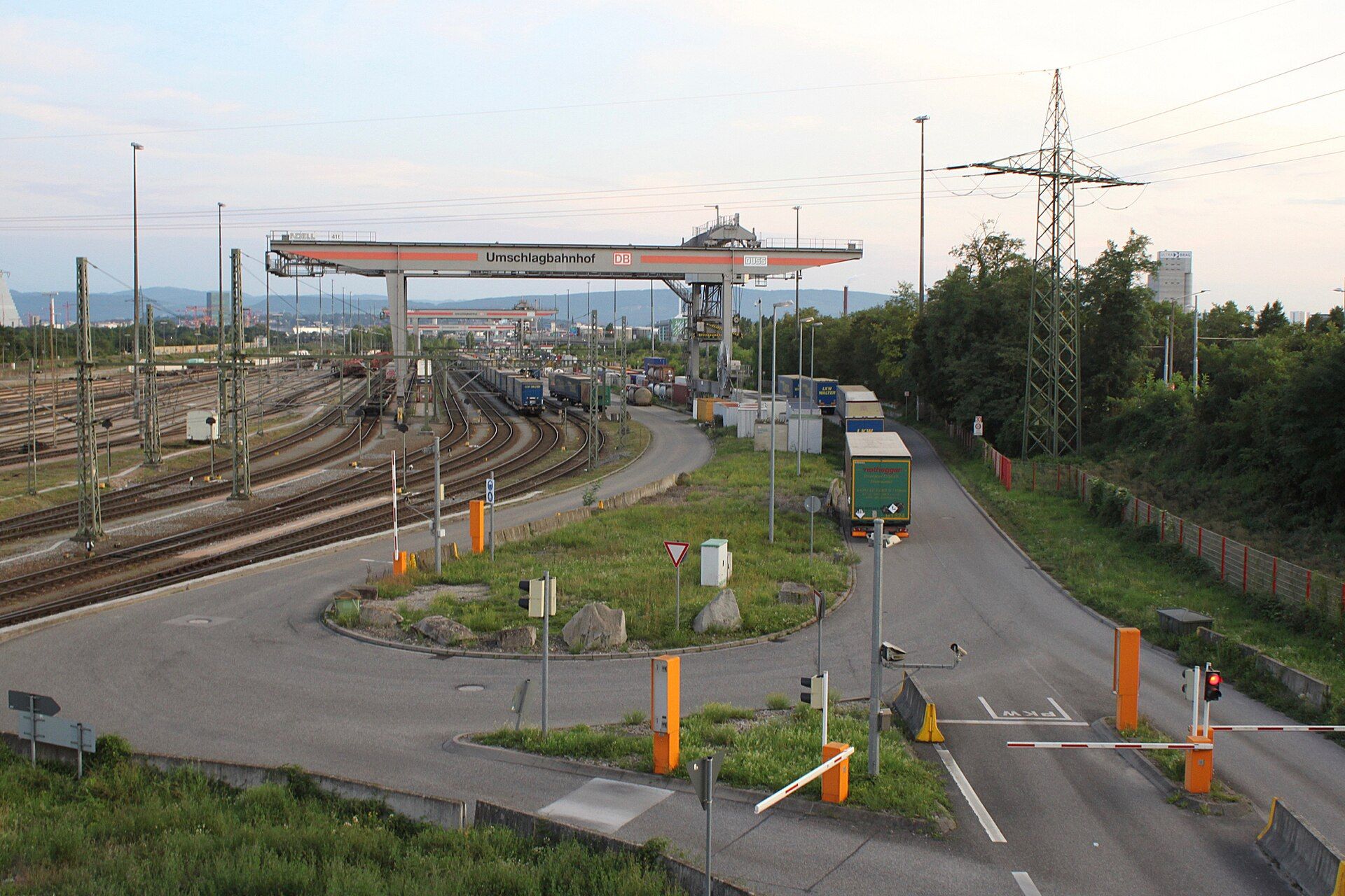 A queue of lorries on the access road of the road–rail transhipment terminal at Weil am Rhein, Germany, with a gantry crane marked Umschlagbahnhof over the tracks, loaded freight trains beside it, parked loading units along the road and red-and-white gate barriers in the foreground