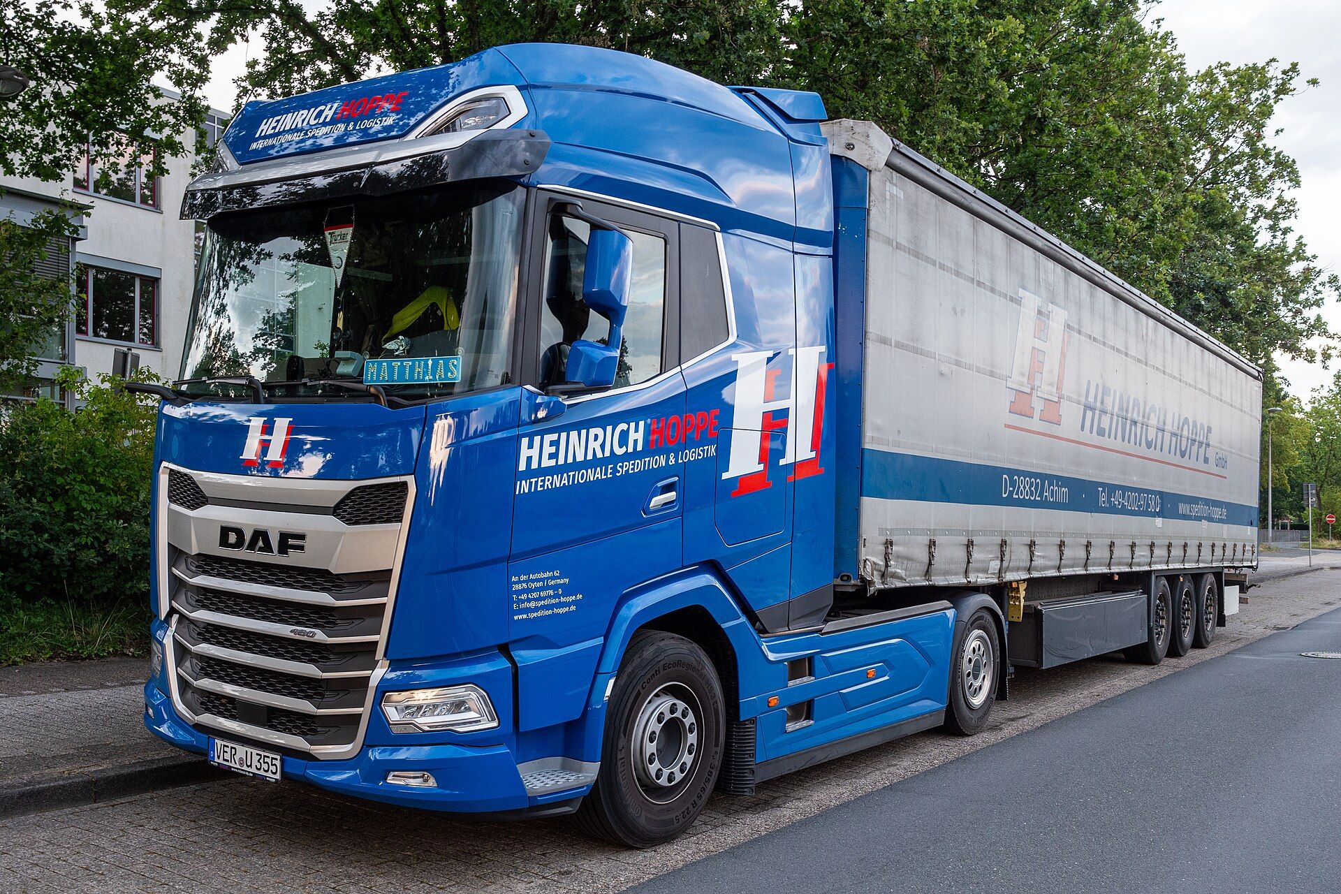 Tracteur routier DAF XF 480 bleu à deux essieux aux couleurs de Heinrich Hoppe, attelé à une semi-remorque à rideaux coulissants à trois essieux, stationné le long du trottoir d'une rue bordée d'arbres