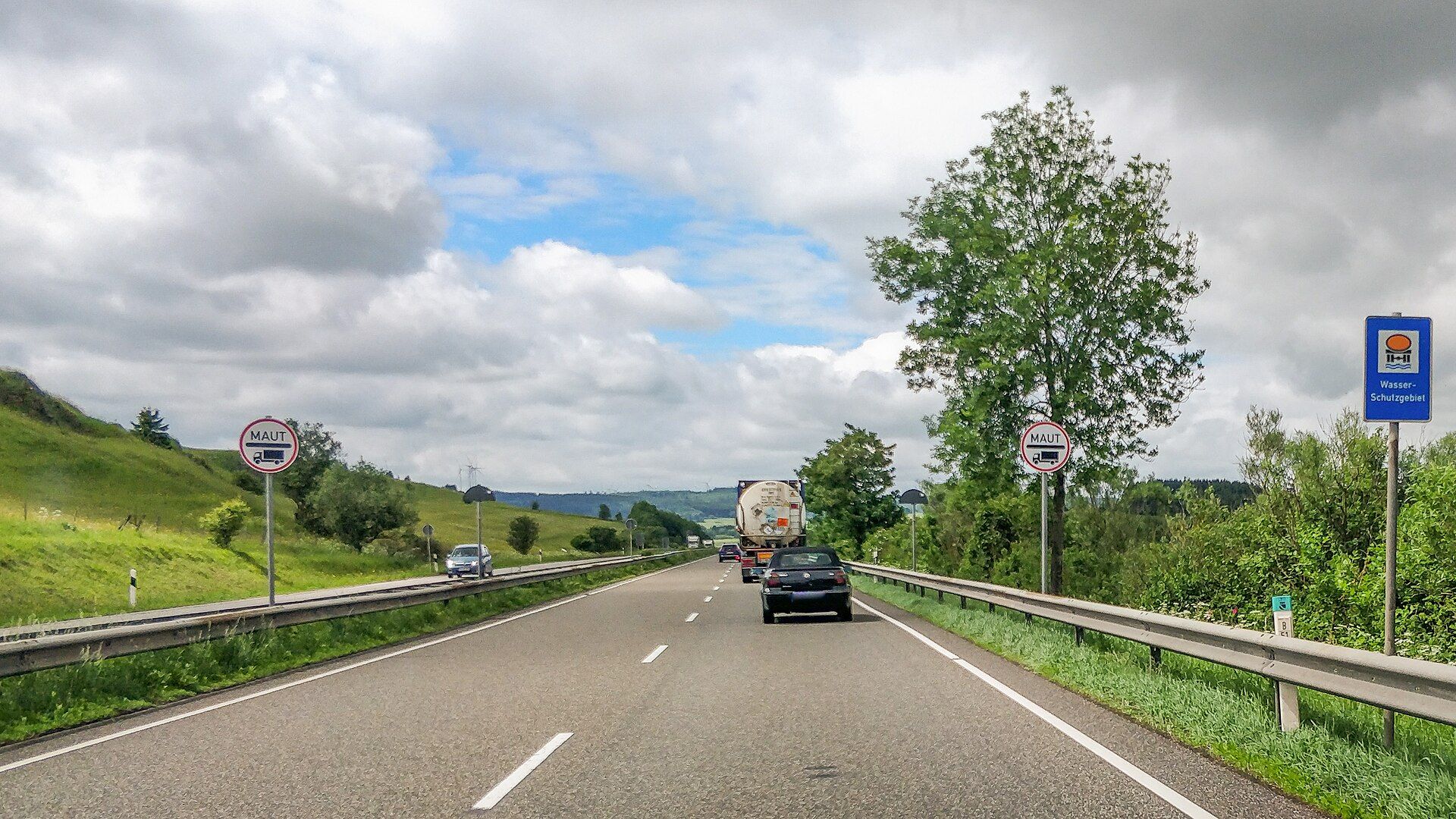 Zwei runde Verkehrszeichen „Maut“ mit Lkw-Symbol zu beiden Seiten der zweistreifigen Richtungsfahrbahn der Bundesstraße 51 bei Dahlem; voraus ein dunkles Cabrio und ein Sattelzug mit Tankcontainer, rechts ein blaues Schild „Wasser-Schutzgebiet“