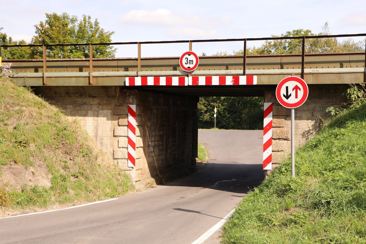 Eisenbahnunterführung mit einem Verkehrszeichen, das die tatsächliche Höhe auf 3 m begrenzt, und rot-weißen Warnmarkierungen an Brückenträger und Widerlagern