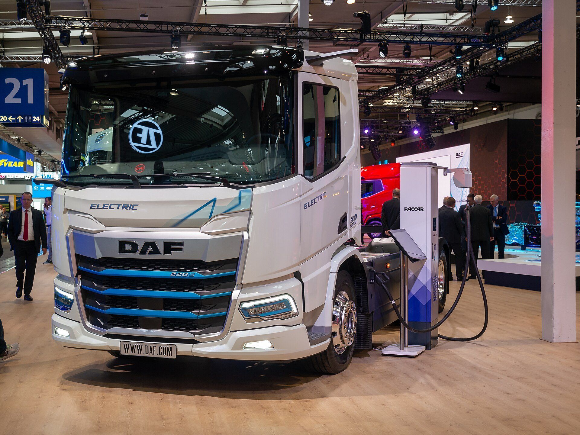 A white DAF XD Electric truck at a trade-fair stand, with a black charging cable running between a PACCAR charging pedestal beside it and the side of the vehicle