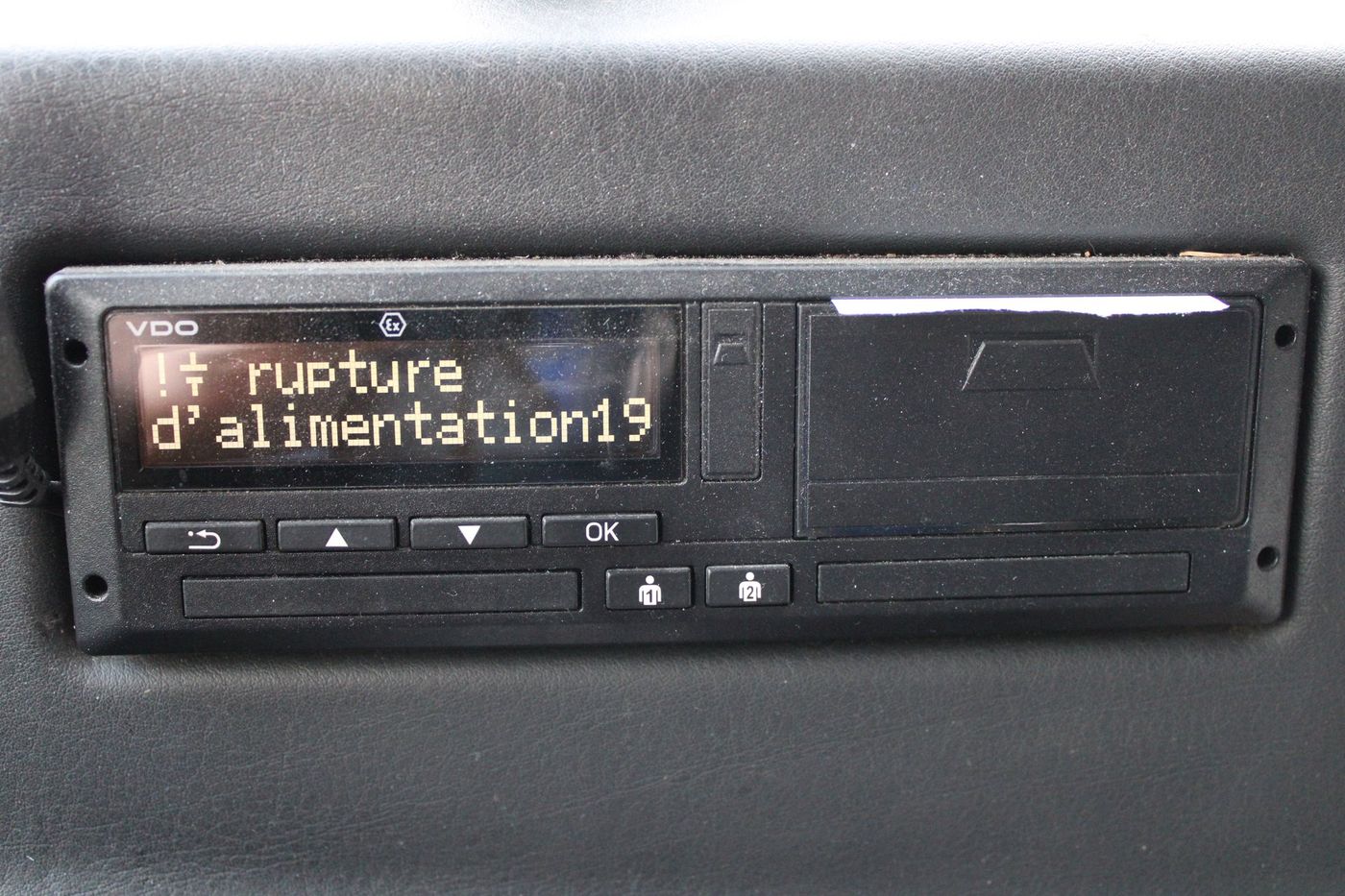 VDO digital tachograph head set into a leather-trimmed panel of a Temsa Tourmalin coach, with two empty card slots, a torn printout strip at the top edge of the printer drawer and a French-language power-supply warning on the display