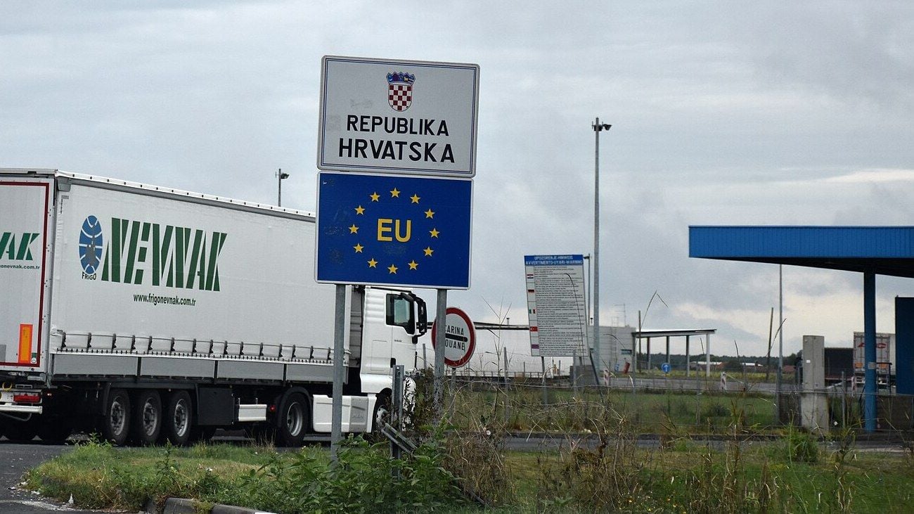White tractor unit and semi-trailer passing the Republika Hrvatska EU border sign at the Bajakovo road crossing in Croatia, with the customs canopy behind