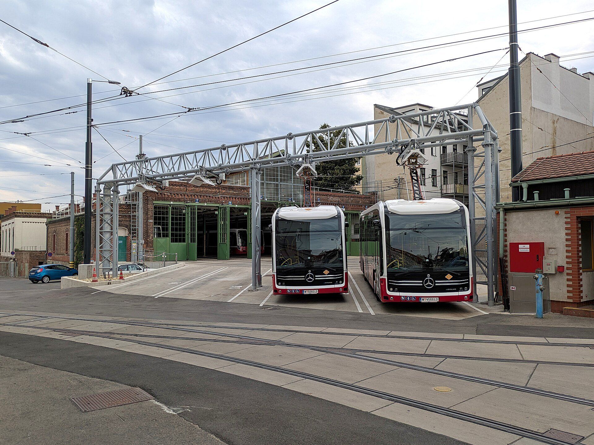 Two Mercedes-Benz eCitaro city buses parked side by side under a steel charging gantry with their roof pantographs raised to it, in front of the doors of a depot hall in Vienna