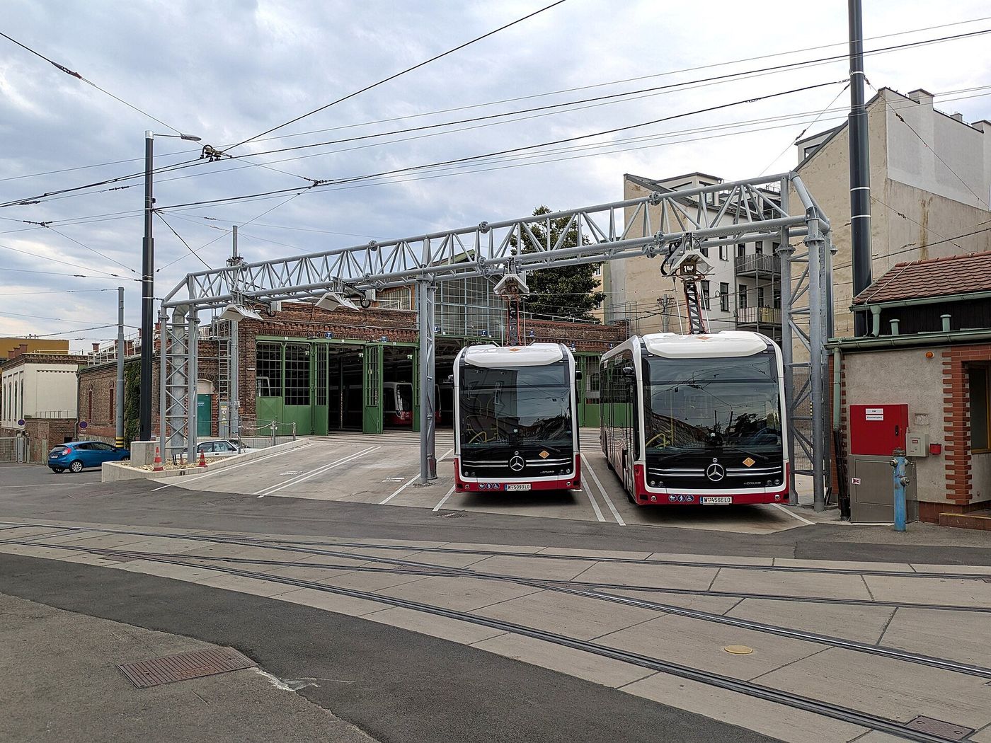 Two Mercedes-Benz eCitaro city buses parked side by side under a steel charging gantry with their roof pantographs raised to it, in front of the doors of a depot hall in Vienna