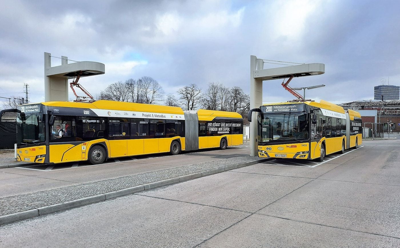Two yellow articulated electric city buses standing under overhead charging masts with pantograph arms at a bus terminal in Berlin