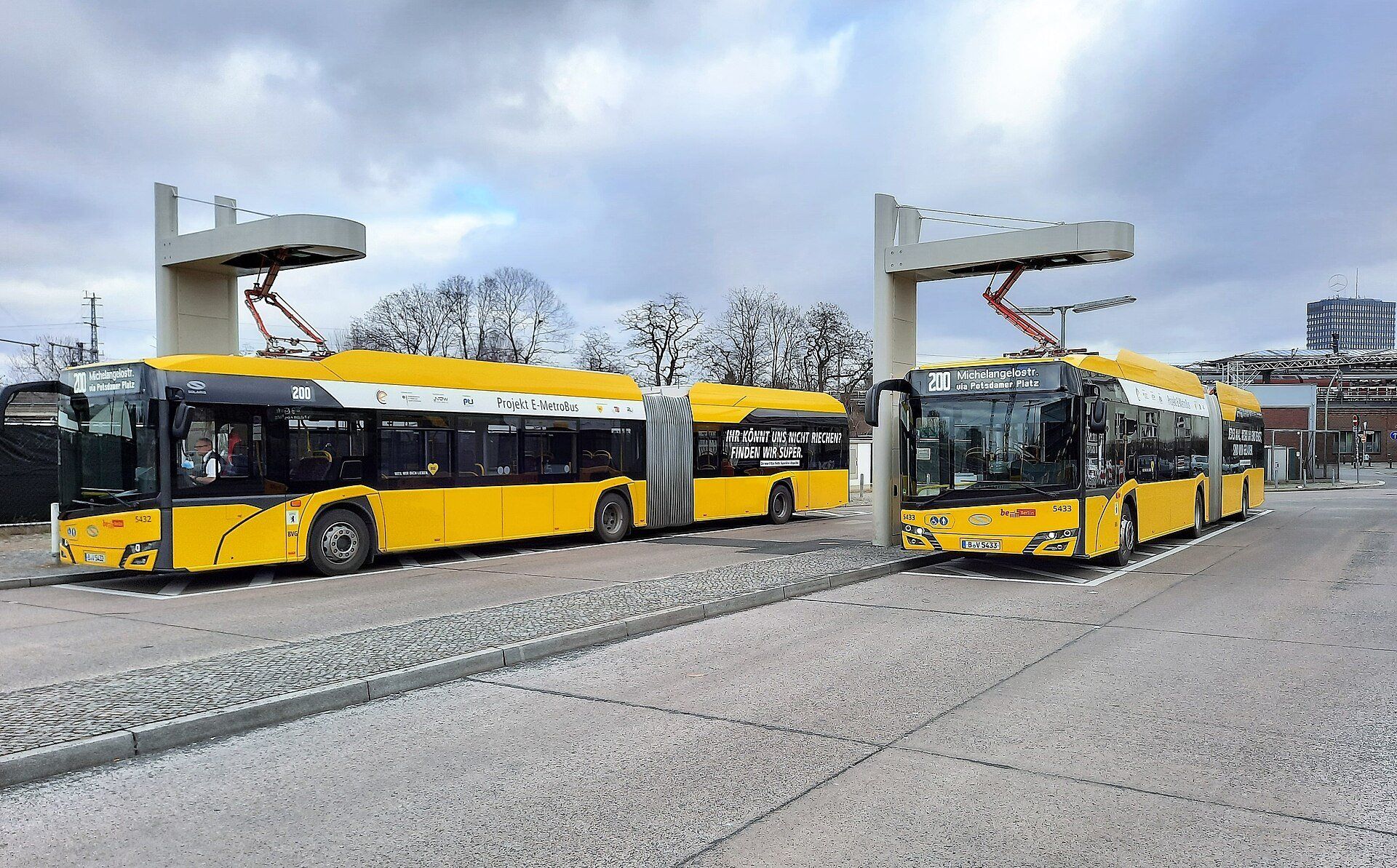 Two yellow articulated electric city buses standing under overhead charging masts with pantograph arms at a bus terminal in Berlin