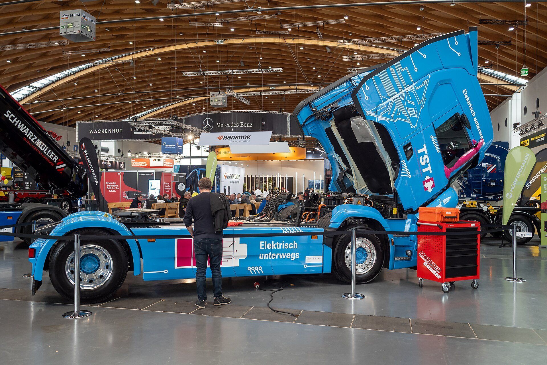 Blue battery-electric tractor unit on display at a trade fair with its cab tilted up, showing the drivetrain bay with orange high-voltage cabling and a red tool trolley beside it
