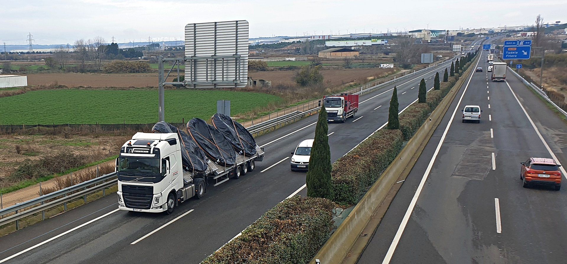 Tractocamión blanco con semirremolque de plataforma y carga cubierta con lonas, seguido de un camión rígido de caja roja, en una carretera de doble calzada en España