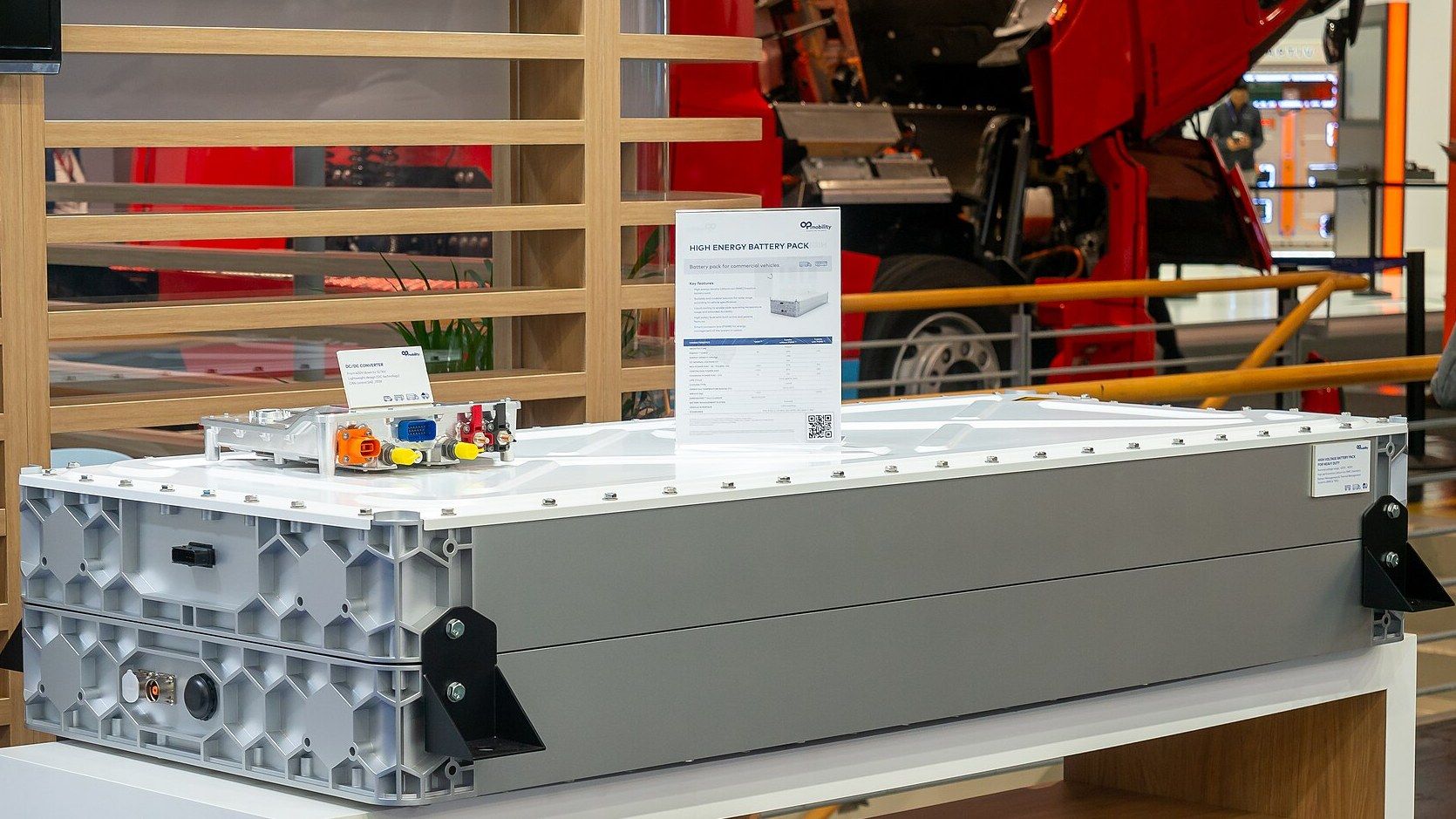 A grey high-energy battery pack for commercial vehicles on a display stand at IAA Transportation 2024 in Hanover, with a DC/DC converter on top, a data sheet beside it and a red truck with its cab tilted forward behind