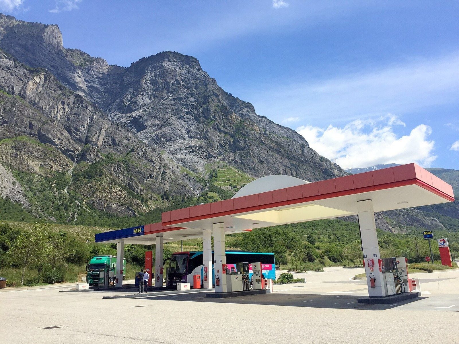 Station-service Total et AS 24 en Savoie en 2016 : un camion Iveco vert et un autocar aux pompes sous l'auvent, au pied d'une paroi rocheuse