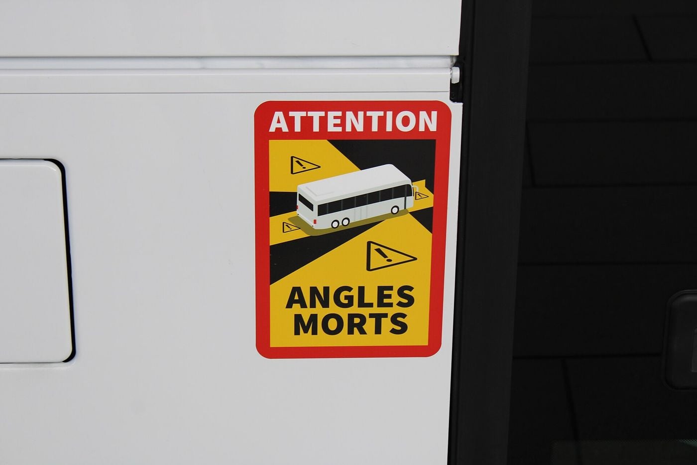 Red and yellow ATTENTION ANGLES MORTS sign with a coach pictogram on the white side panel of a bus, next to a dark window