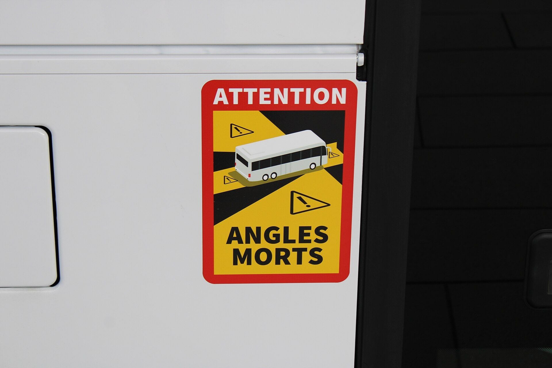 Red and yellow ATTENTION ANGLES MORTS sign with a coach pictogram on the white side panel of a bus, next to a dark window