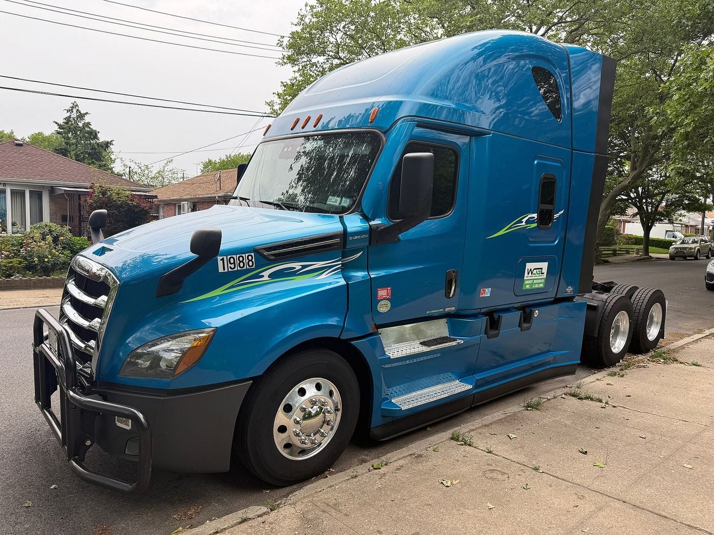 Blue Freightliner Cascadia sleeper tractor with a grille guard and tandem drive axles, parked without a trailer at the kerb of a residential street