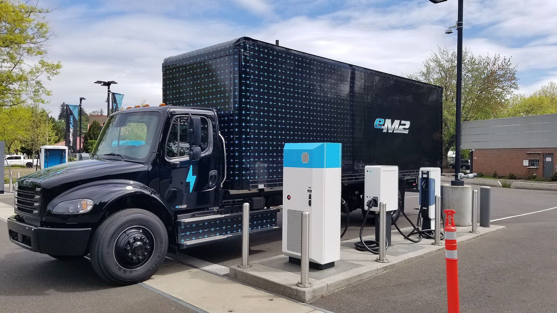 Black Freightliner eM2 box truck with a wrap of small blue crosses and eM2 lettering, parked beside white charging posts at a charging site in Portland, Oregon, photographed in 2024