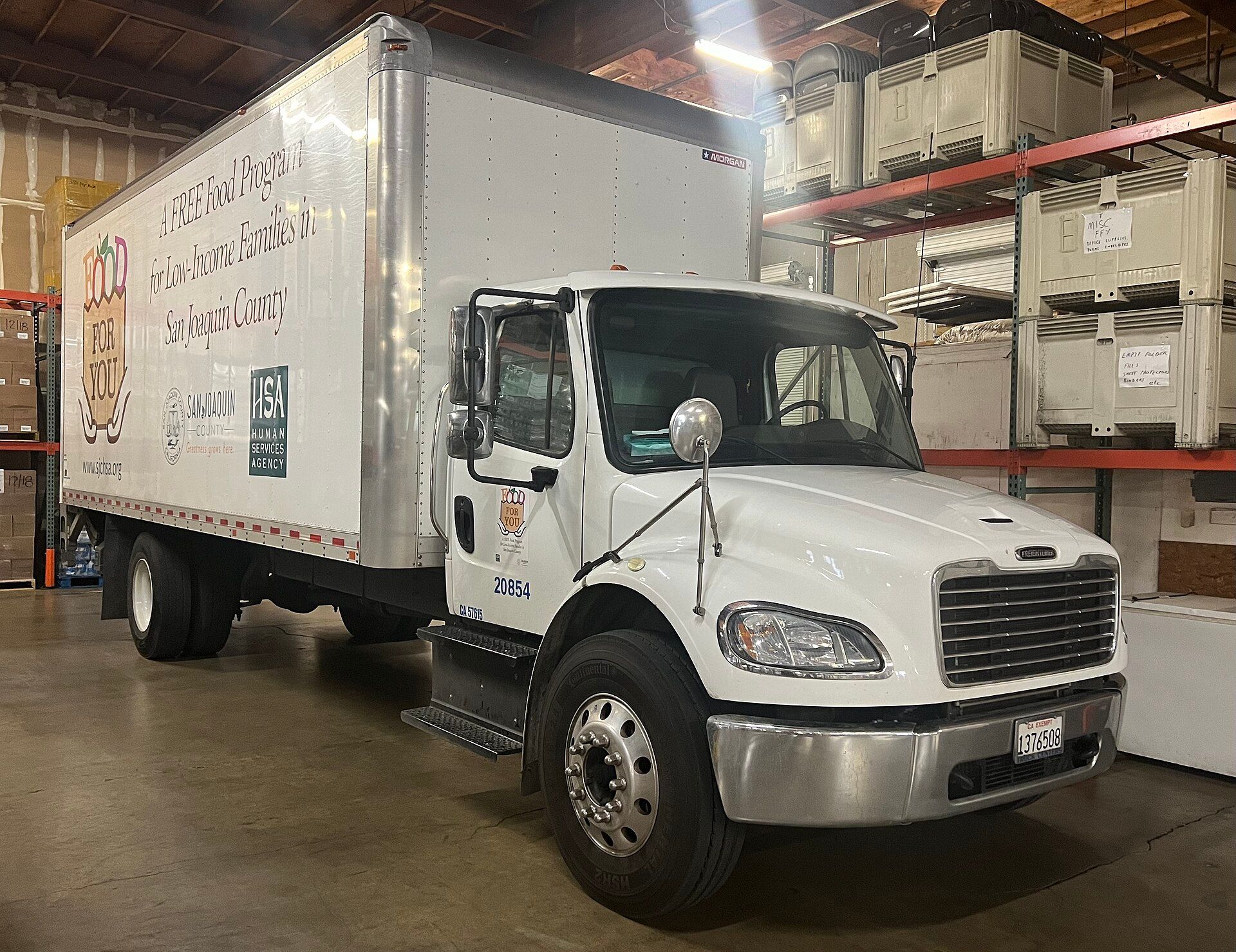 White Freightliner M2 day-cab truck with a box body, one rear axle, a chrome bumper and a convex hood-mounted mirror, parked inside a warehouse beside shelves of storage bins