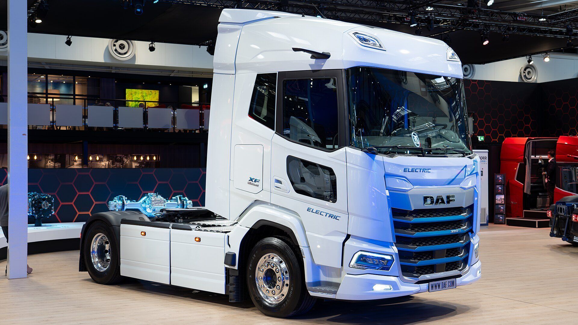A white DAF XF Electric tractor unit with blue trim on a trade-fair stand, Electric lettering on the front and cab side, and a displayed axle unit behind the cab