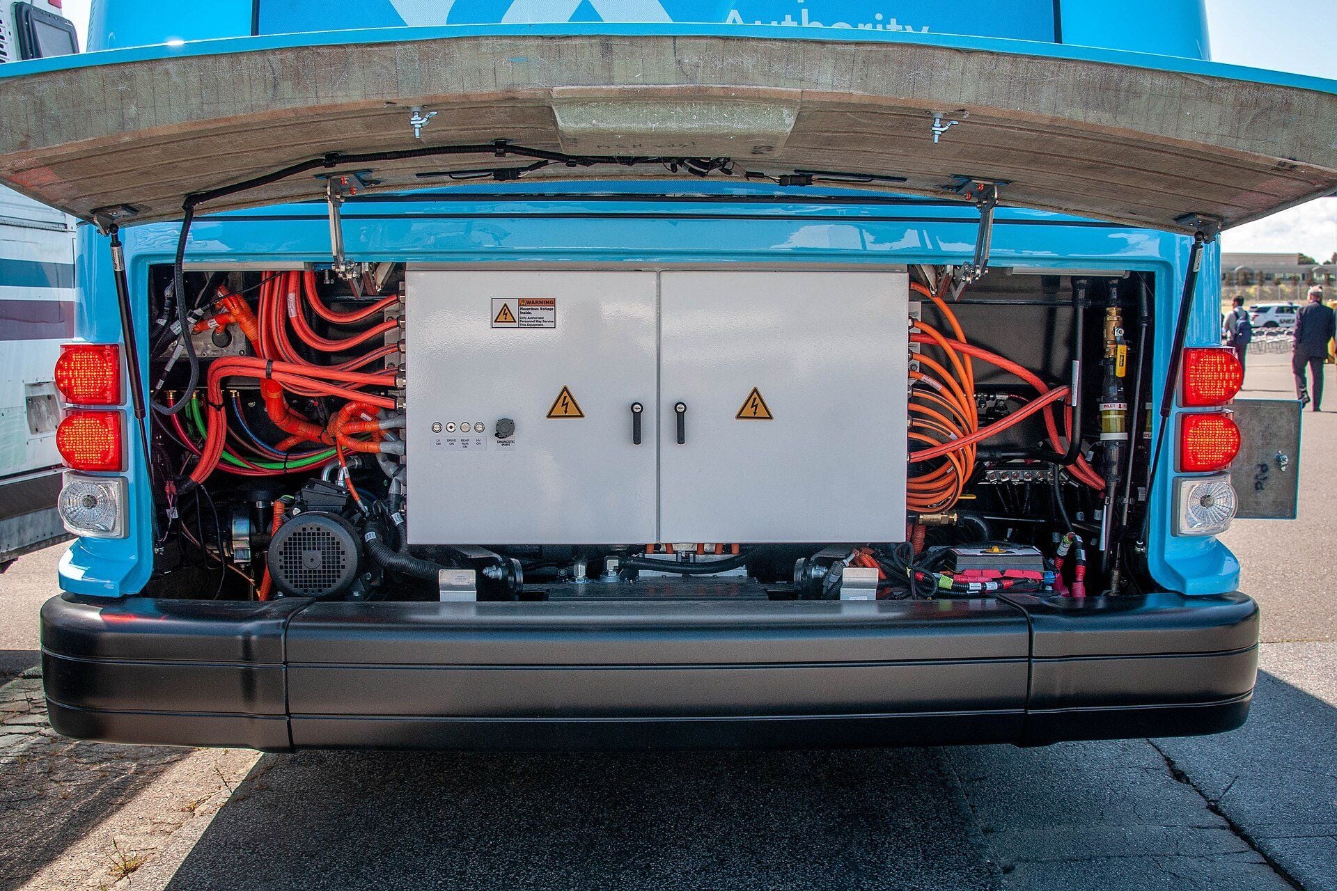 Open rear compartment of an electric city bus with bundles of orange high-voltage cables on both sides of a grey cabinet carrying hazardous-voltage warning labels
