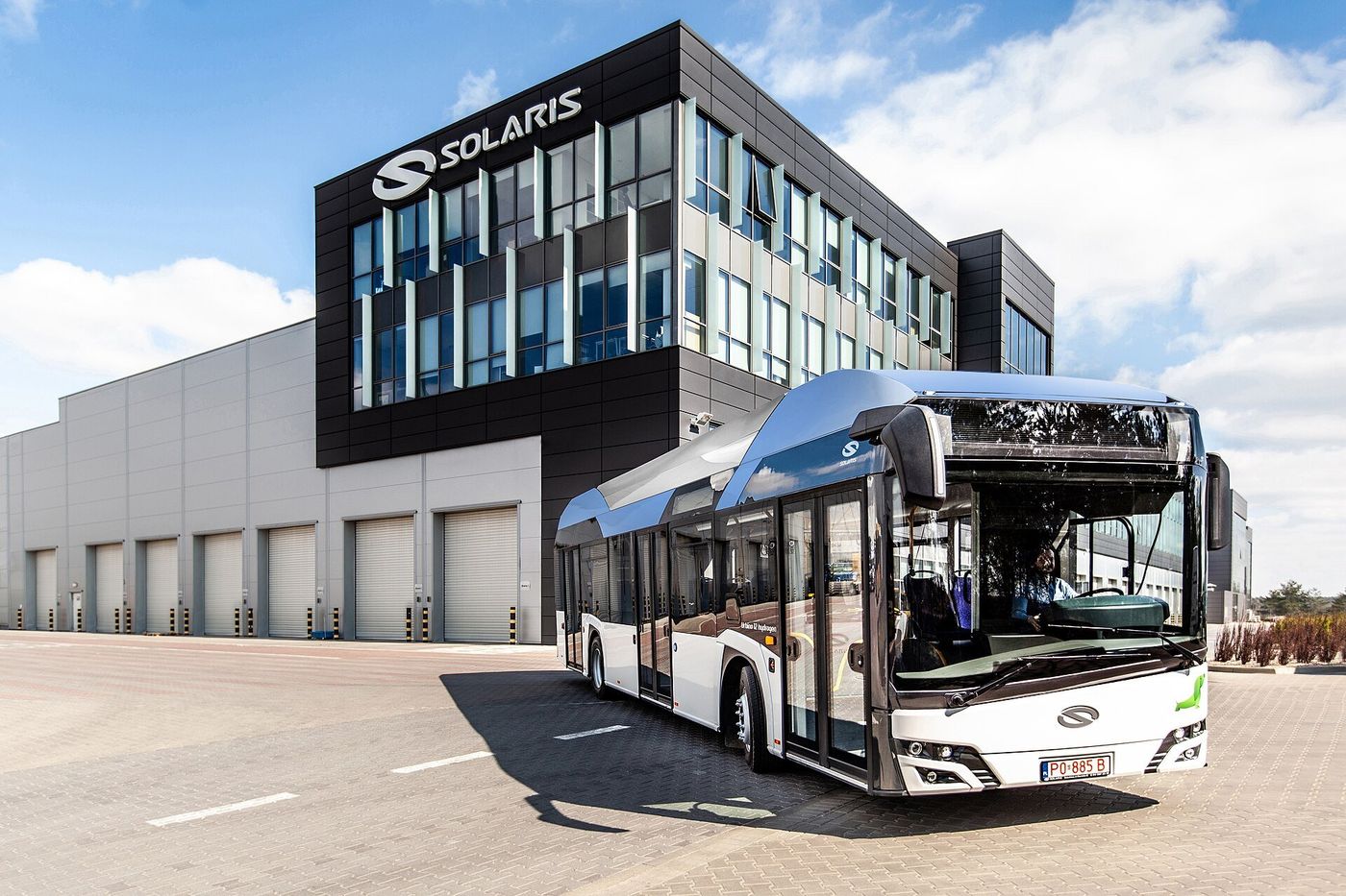 A white Solaris Urbino 12 hydrogen city bus with a grey-blue roof section parked in front of a building carrying the Solaris name, seen from the front right on a sunny day