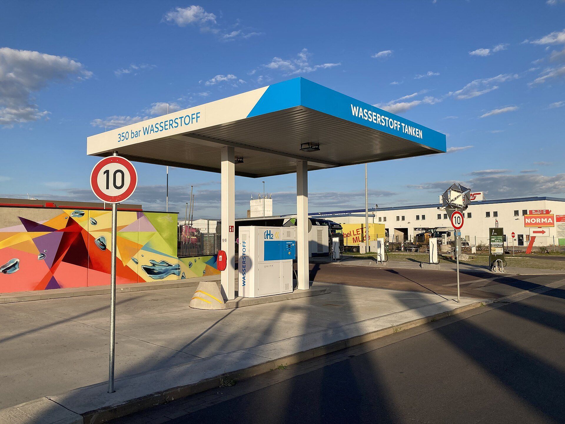 A 350 bar hydrogen dispenser under a blue-and-white canopy marked Wasserstoff tanken at a filling station in Magdeburg, Germany, with parked lorries in the background