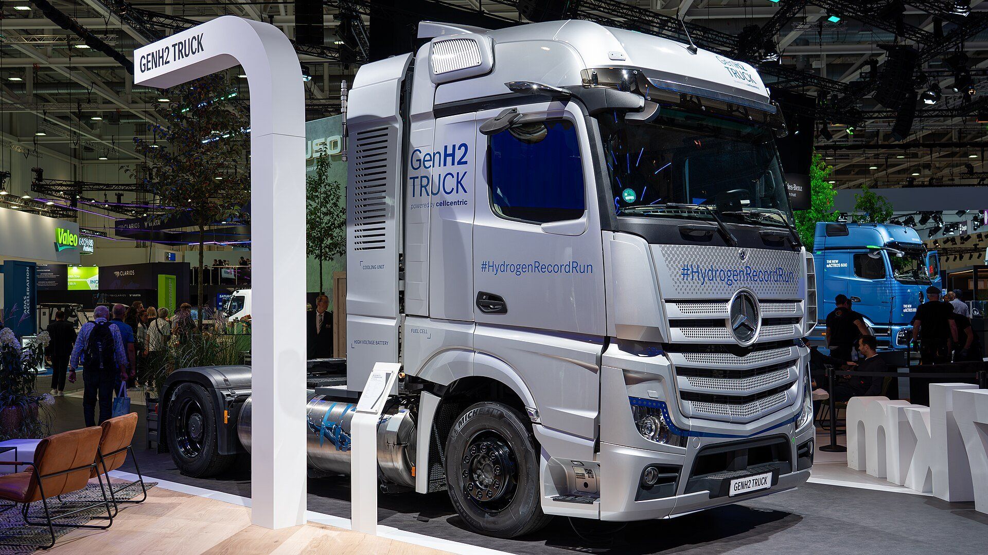 A silver Mercedes-Benz GenH2 Truck tractor unit on a trade-fair stand, with Fuel Cell and High Voltage Battery labels on the panel behind the cab and a cylindrical tank between the axles