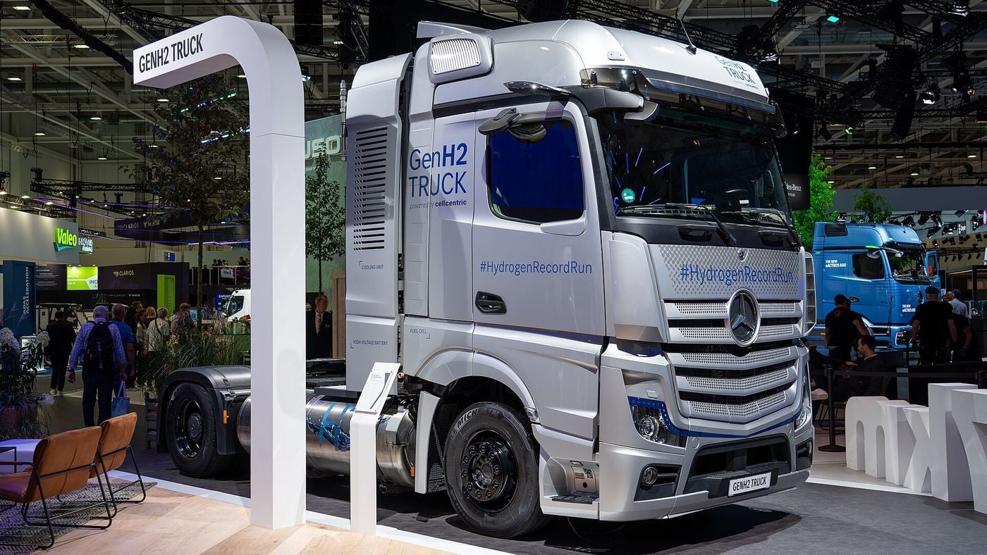 A silver Mercedes-Benz GenH2 Truck tractor unit on a trade-fair stand, with Fuel Cell and High Voltage Battery labels on the panel behind the cab and a cylindrical tank between the axles