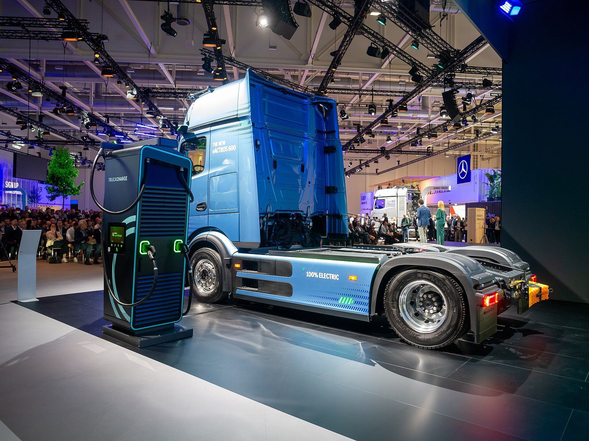 A blue Mercedes-Benz eActros 600 tractor unit on a trade-fair stand, parked beside a charging cabinet labelled TRUCKCHARGE, with 100% ELECTRIC printed on its side fairing