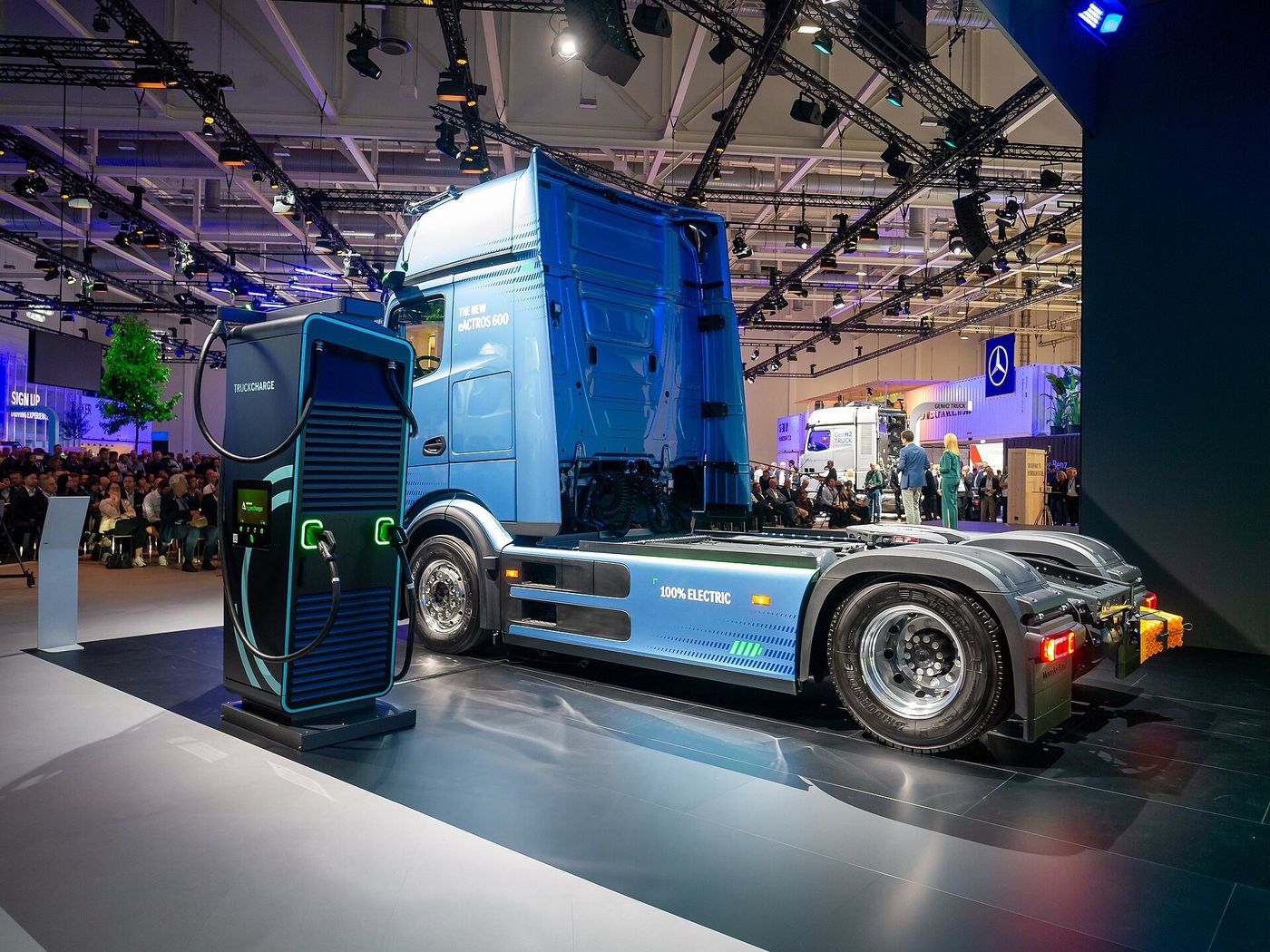 A blue Mercedes-Benz eActros 600 tractor unit on a trade-fair stand, parked beside a charging cabinet labelled TRUCKCHARGE, with 100% ELECTRIC printed on its side fairing
