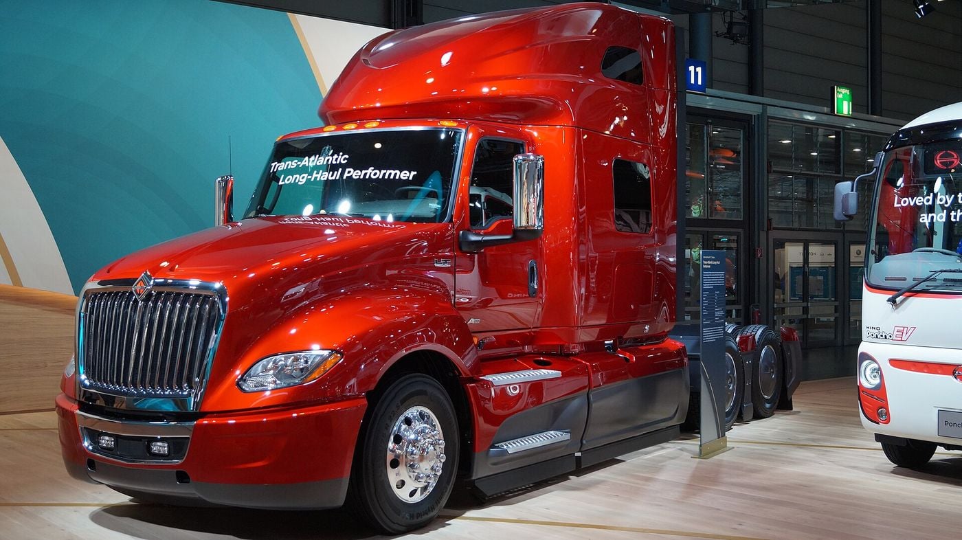 Red International LT625 sleeper tractor with a high roof, a chrome vertical-bar grille and two rear axles, shown without a trailer on a stand at the IAA 2018 show, with 'Trans-Atlantic Long-Haul Performer' lettered on the windshield