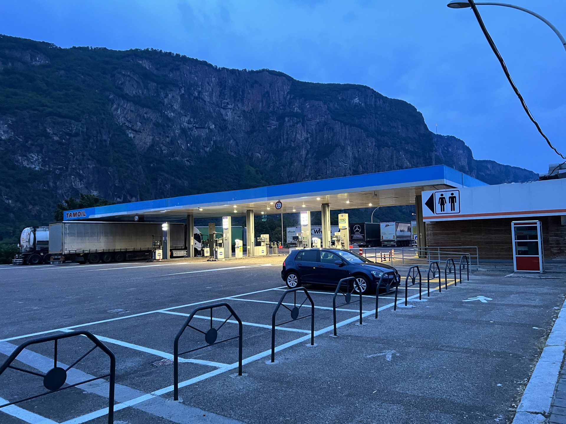 Distributore Tamoil dell'area di servizio «Laimburg Ost» sull'autostrada A22 del Brennero a Vadena, in Alto Adige, all'imbrunire: sotto la pensilina illuminata un'autocisterna e un autoarticolato telonato alle pompe, altri camion fermi sullo sfondo, un'auto scura nel piazzale e dietro la parete rocciosa della montagna