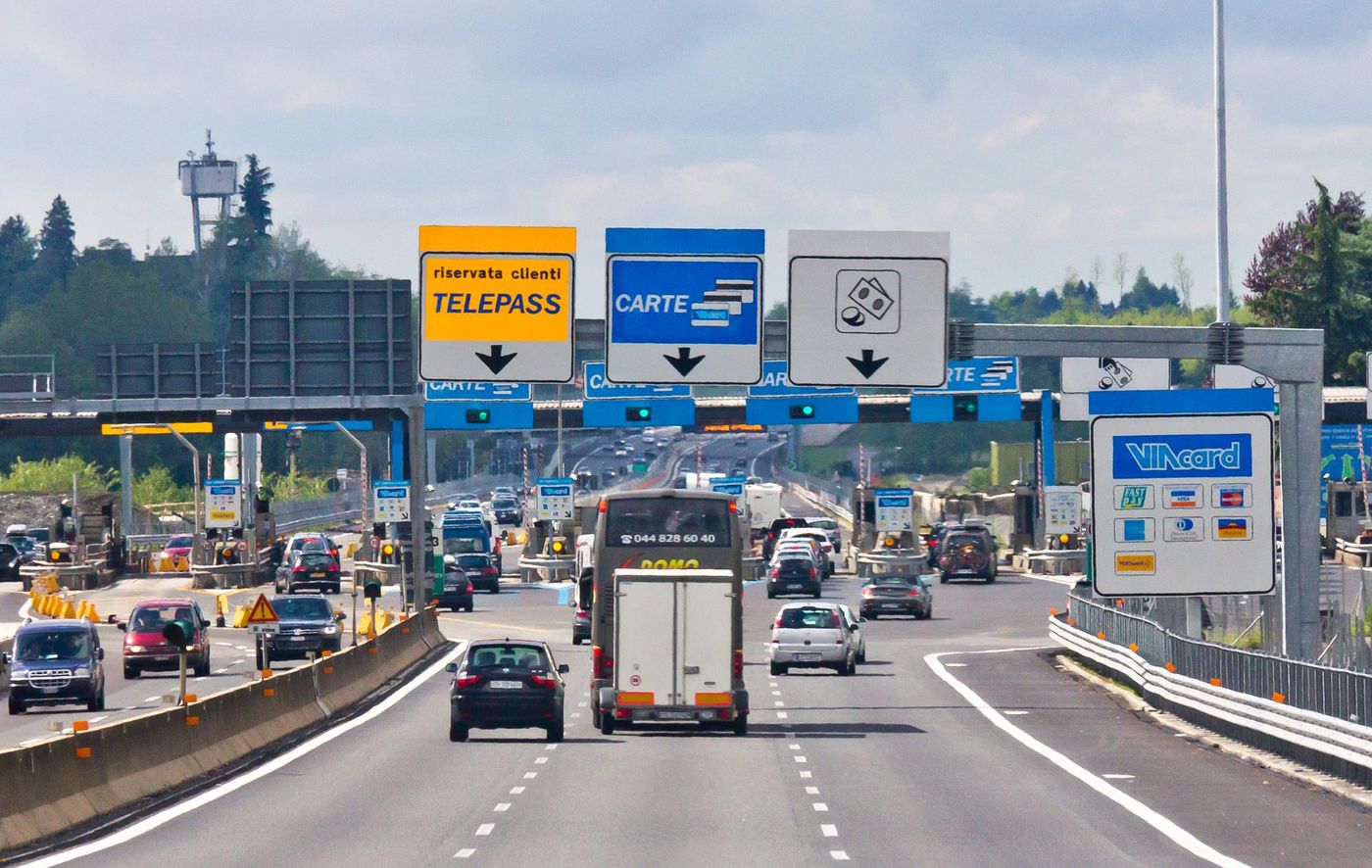Un pullman che traina un rimorchio chiuso, tra altre auto, si avvicina alla stazione di pedaggio di Como Grandate sull'autostrada A9 in direzione sud; sopra le corsie i cartelli «riservata clienti TELEPASS» su fondo giallo, «CARTE» su fondo blu e quello bianco con monete e banconote, a destra il cartello Viacard con i loghi delle carte accettate