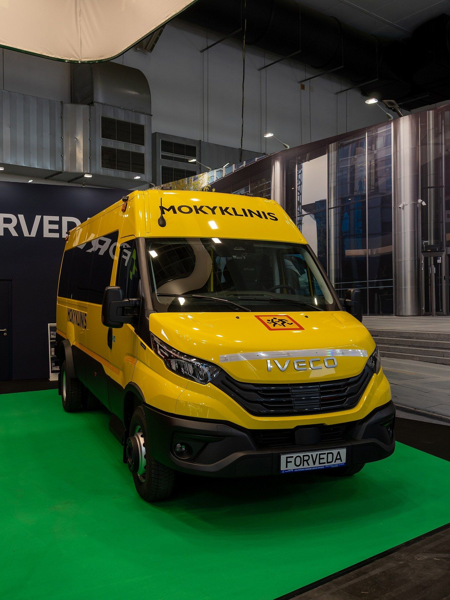 Yellow minibus on an Iveco Daily van base with a school-transport sign on the bonnet, the word MOKYKLINIS above the windscreen and a FORVEDA plate, on a stand at Busworld Europe 2025 in Brussels