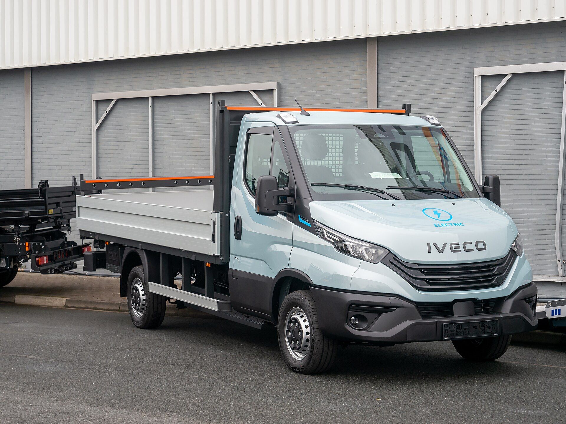 Pale blue battery-electric Iveco eDaily single cab with an ELECTRIC emblem on the bonnet and an aluminium dropside body, parked outside a hall at IAA Transportation 2024 in Hanover