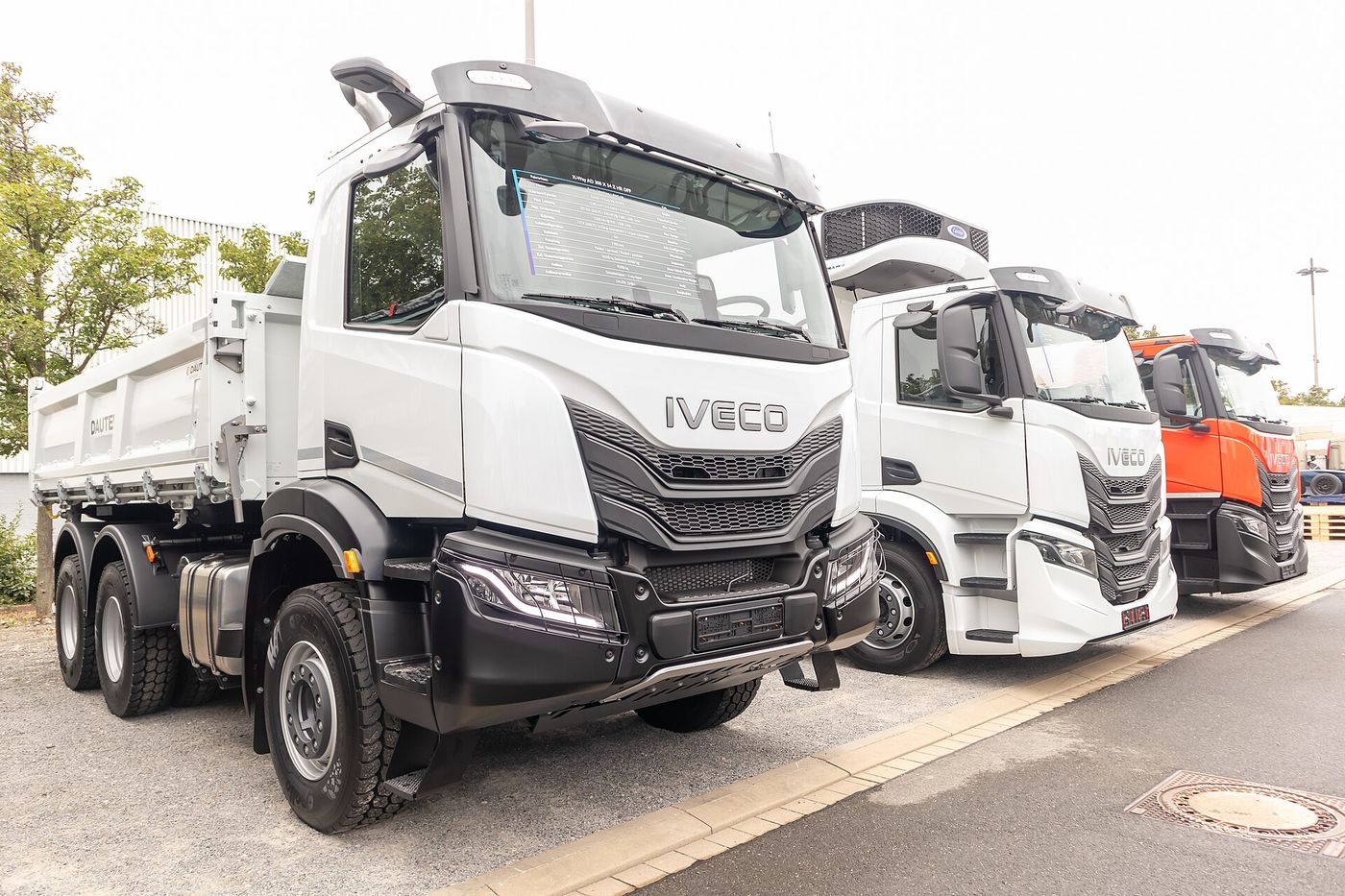 White Iveco X-Way 6x4 tipper with the short day cab and a black front bumper, front three-quarter view outdoors at IAA Transportation 2024 in Hanover, with two more Iveco trucks parked behind it