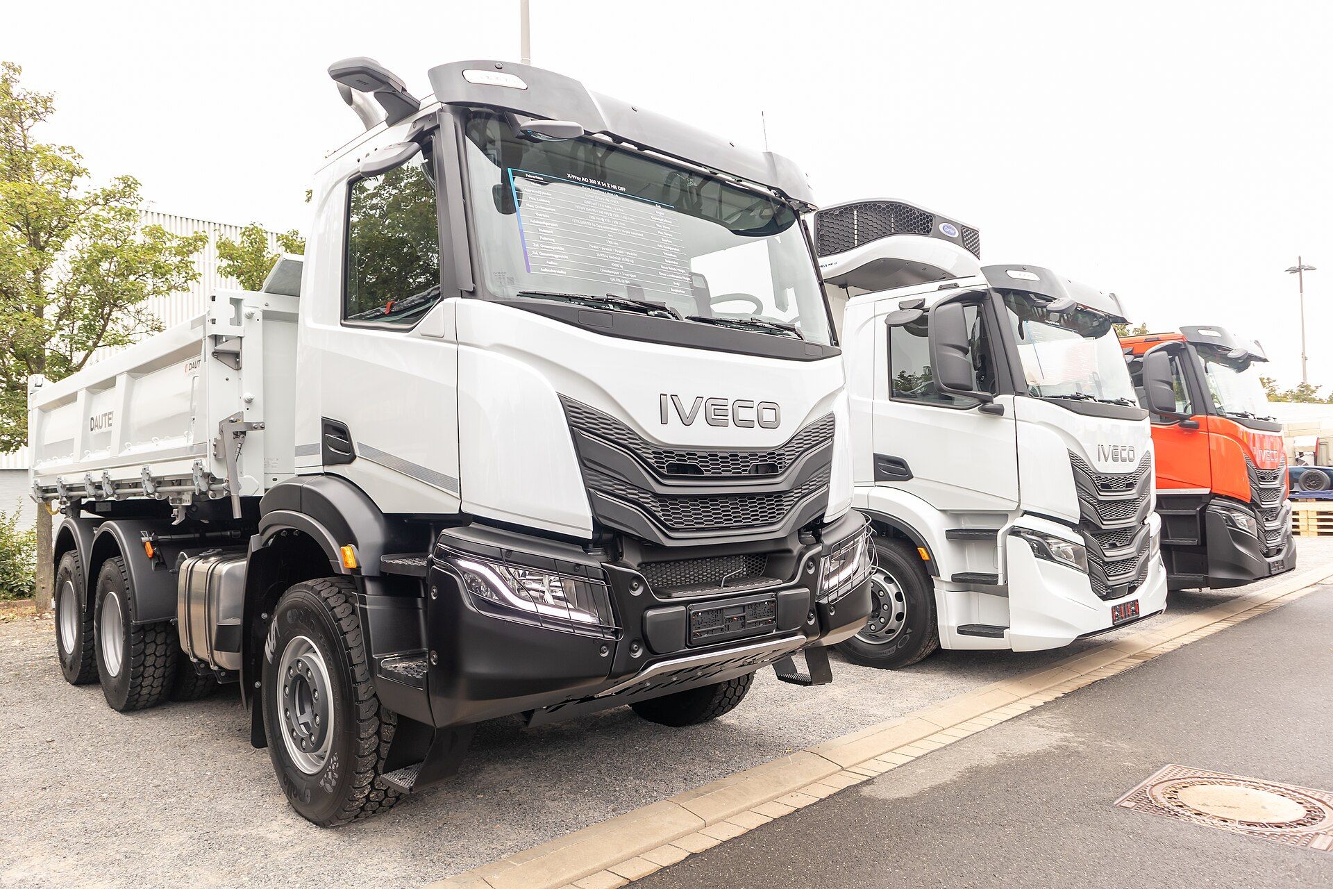 White Iveco X-Way 6x4 tipper with the short day cab and a black front bumper, front three-quarter view outdoors at IAA Transportation 2024 in Hanover, with two more Iveco trucks parked behind it