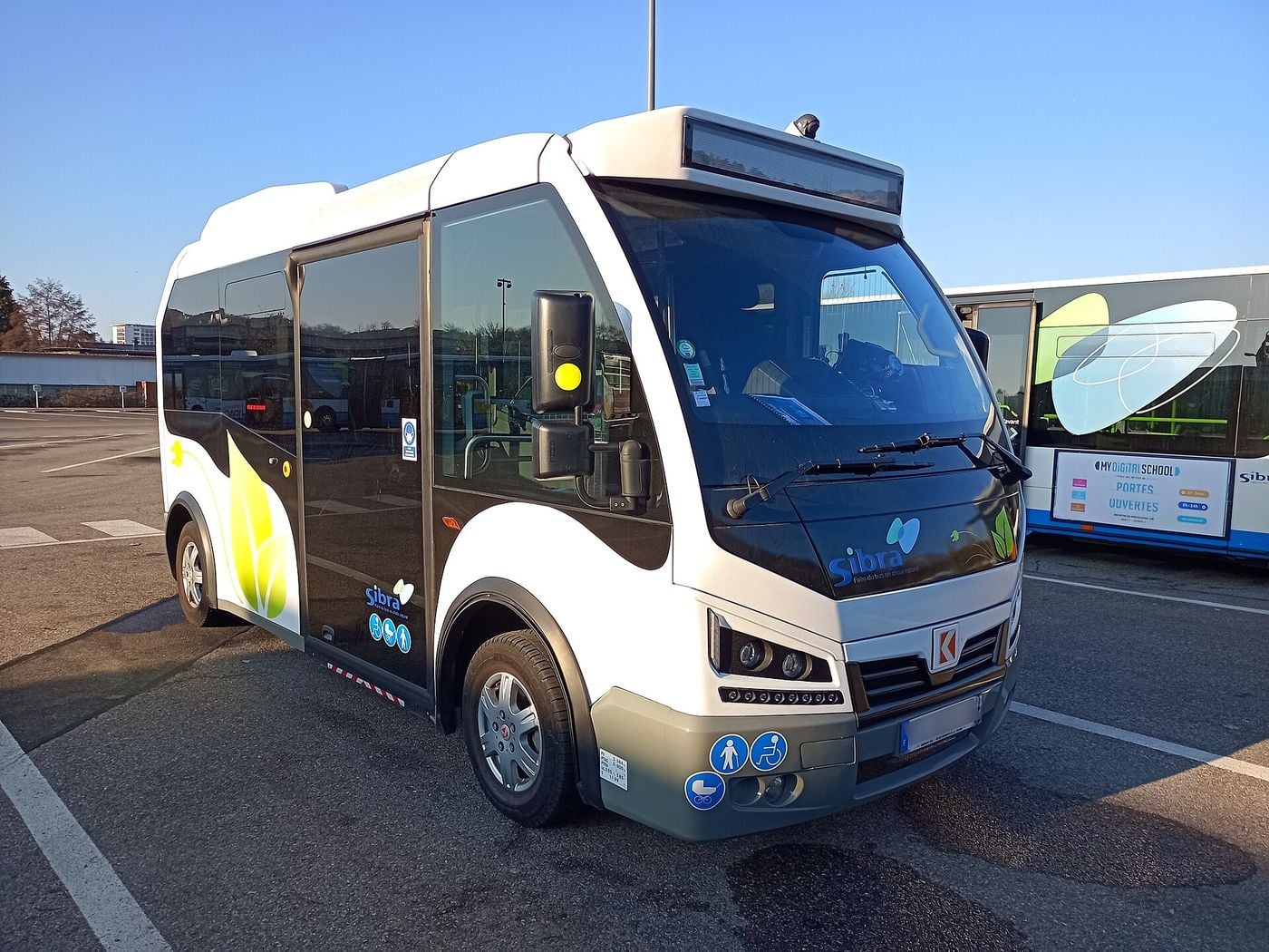 White two-axle Karsan e-JEST battery-electric minibus with a black window band, green leaf graphics and small SIBRA logos, seen from the front right on a depot car park in Annecy, with a city bus parked beside it