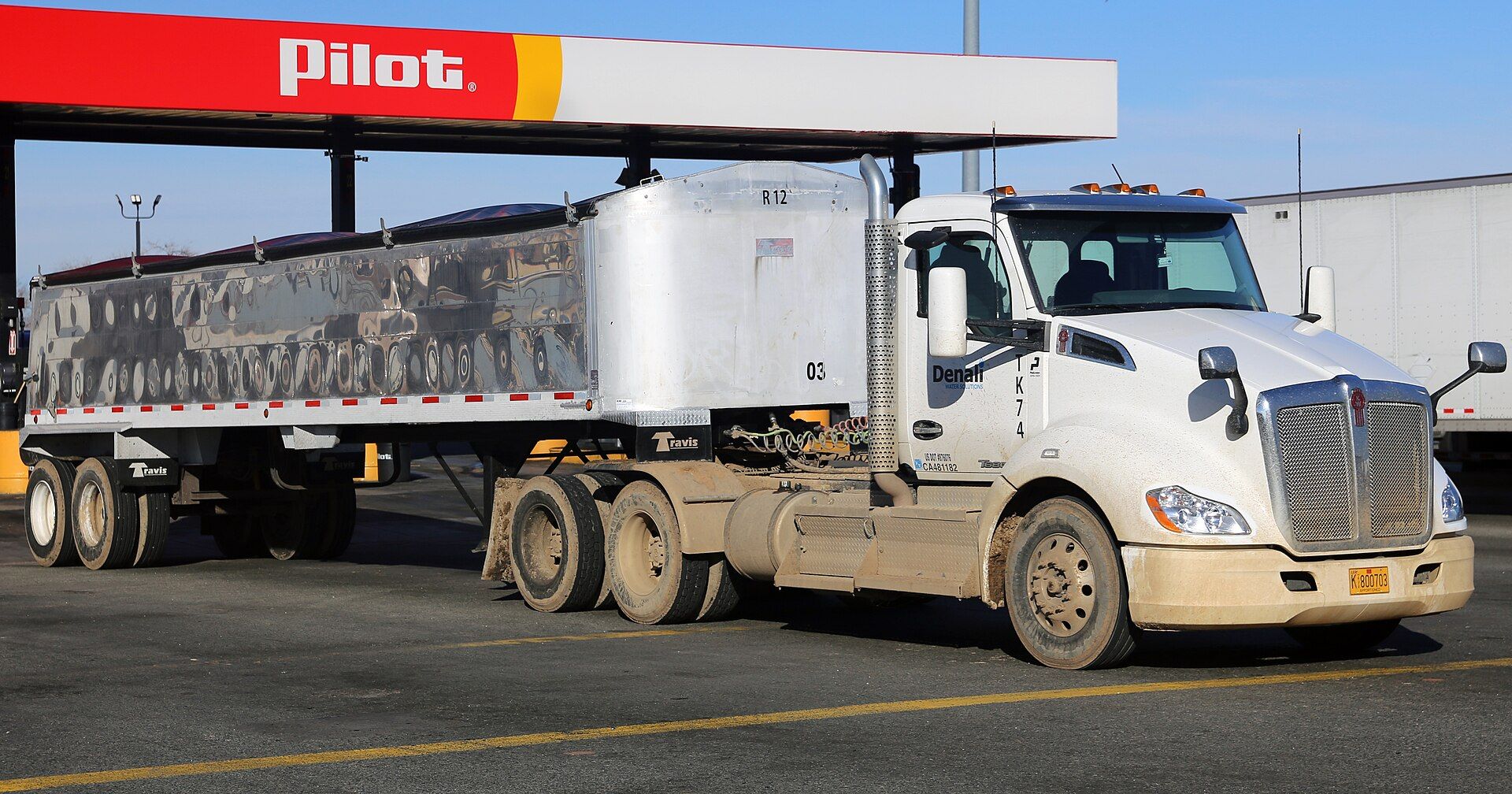 White Kenworth T680 day-cab tractor photographed in 2016, with a wide chrome grille, a vertical exhaust stack and two rear axles, pulling a polished aluminium dump trailer in front of a truck-stop canopy