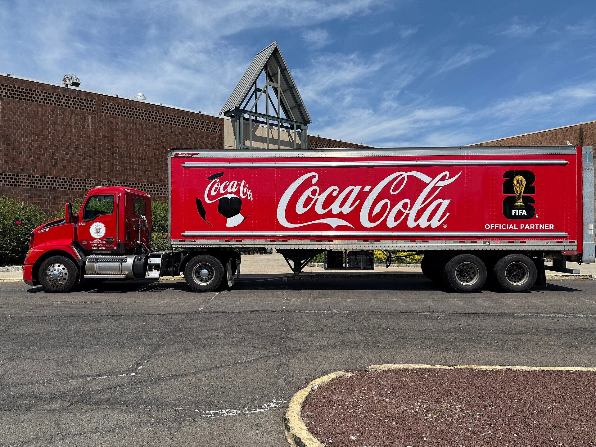 Red Kenworth T680 day-cab tractor with one rear axle and a T680 badge on the door, pulling a red Coca-Cola semi-trailer, seen side-on in a shopping-centre car park