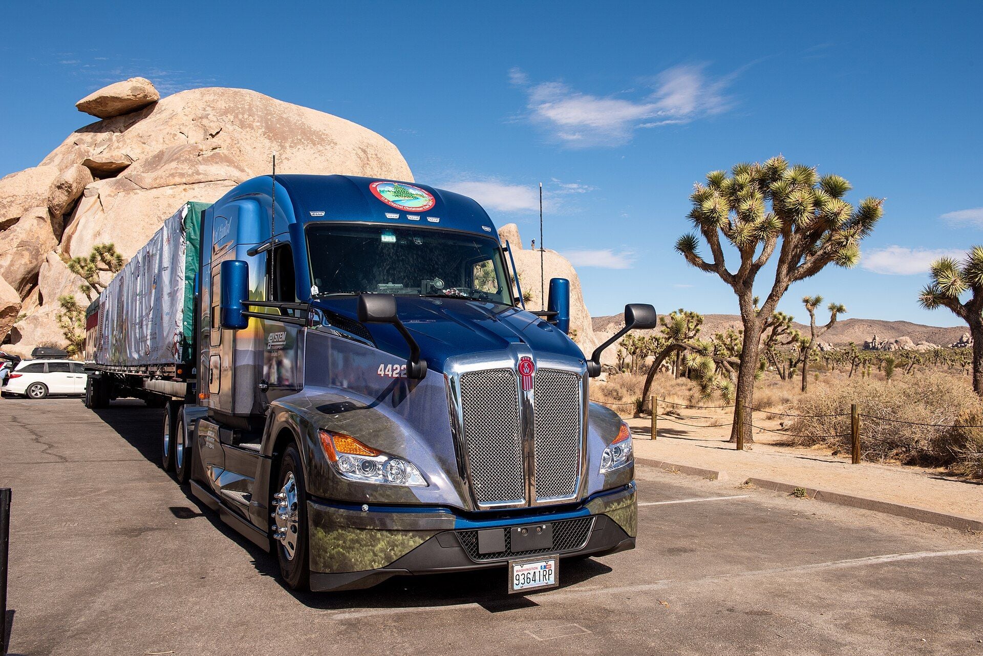 Trattore stradale Kenworth T680 blu dell'attuale generazione con cabina con cuccetta, calandra cromata a rete e grafiche U.S. Capitol Christmas Tree applicate in pellicola, con un semirimorchio pianale dal carico telonato, parcheggiato accanto a massi di granito e alberi di Giosuè nel Parco nazionale di Joshua Tree
