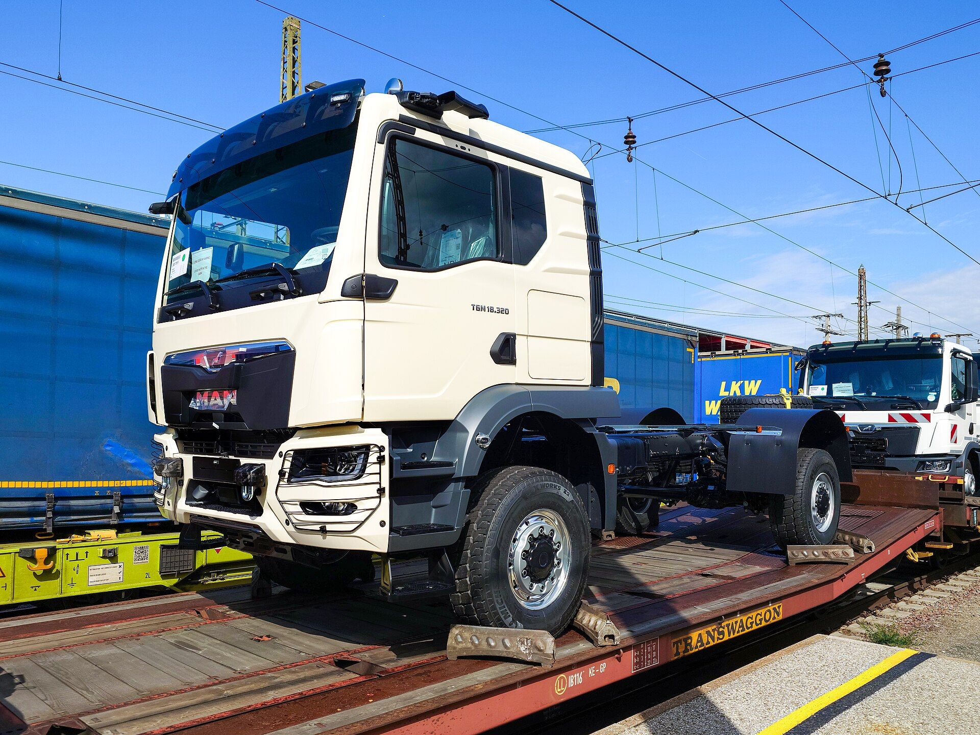 Factory-new cream MAN TGM 18.320 chassis cab with a long cab, a high-mounted frame and large single tyres, carried on a rail wagon at Wels main station