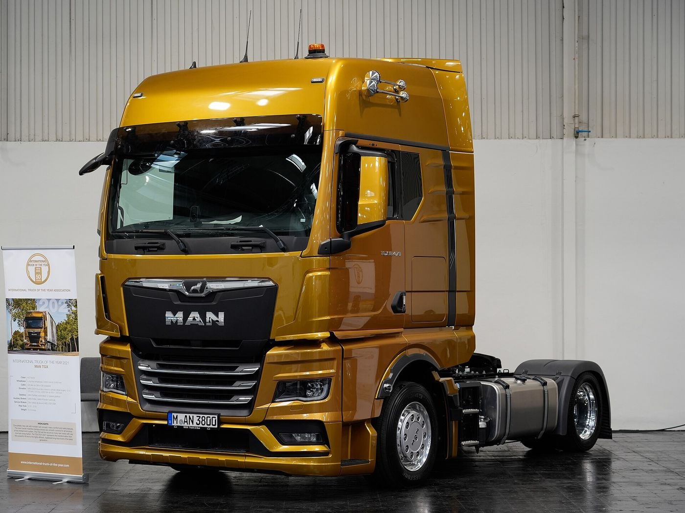 Gold current-generation MAN TGX 18.470 two-axle tractor unit with a high cab, beside an International Truck of the Year banner at a trade-fair display