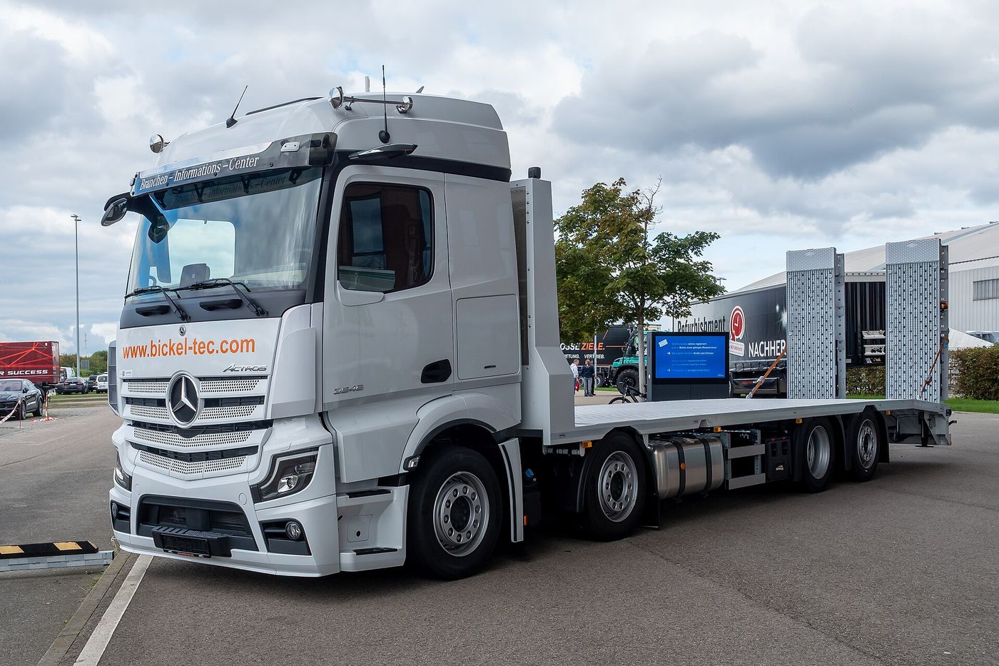 White Mercedes-Benz Actros three-axle rigid with a plant-transporter body and folded loading ramps, seen from the front left on the outdoor display area of Nufam 2025 in Rheinstetten