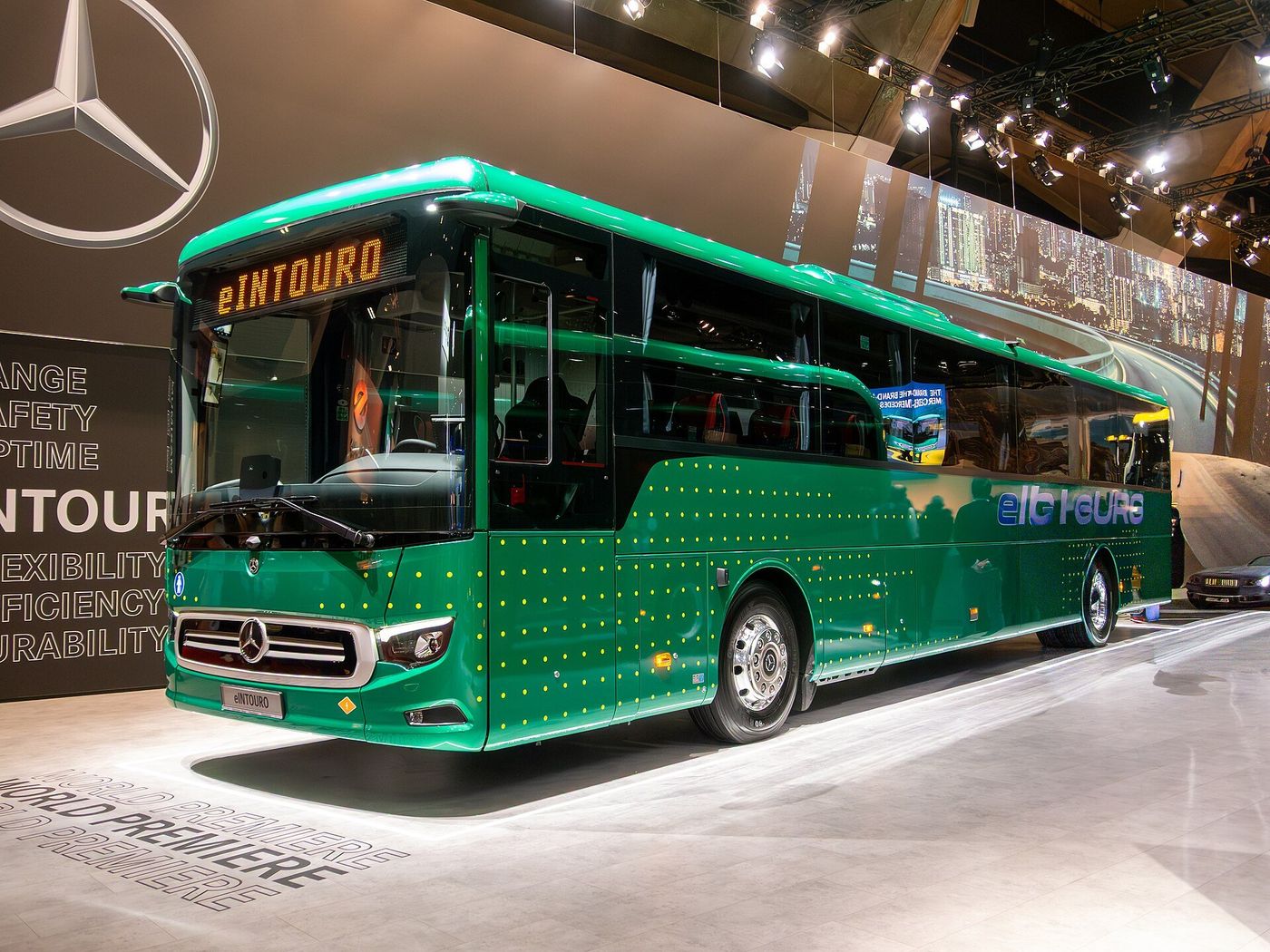 Green Mercedes-Benz eIntouro bus on a display stand at Busworld Europe 2025 in Brussels, seen from the front and left side, with the name eINTOURO on the destination sign