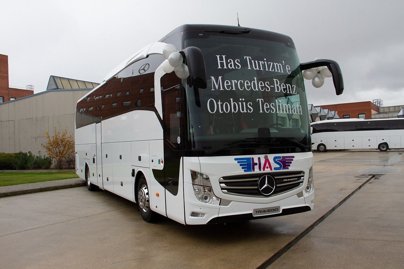 White Mercedes-Benz Travego two-axle touring coach with a TRAVEGO grille badge and a Turkish delivery message on the windscreen, parked on a wet forecourt in front of factory buildings
