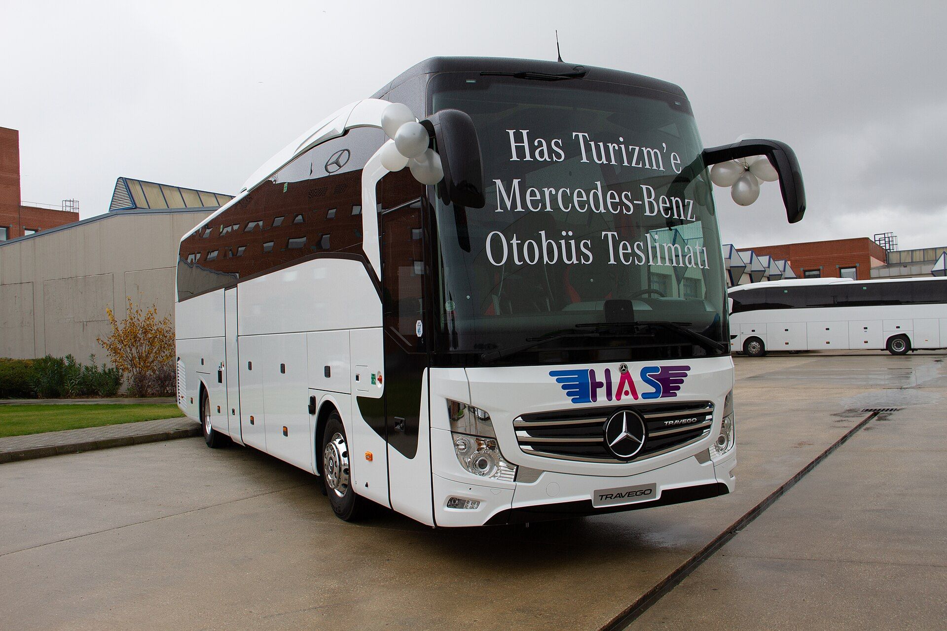 White Mercedes-Benz Travego two-axle touring coach with a TRAVEGO grille badge and a Turkish delivery message on the windscreen, parked on a wet forecourt in front of factory buildings