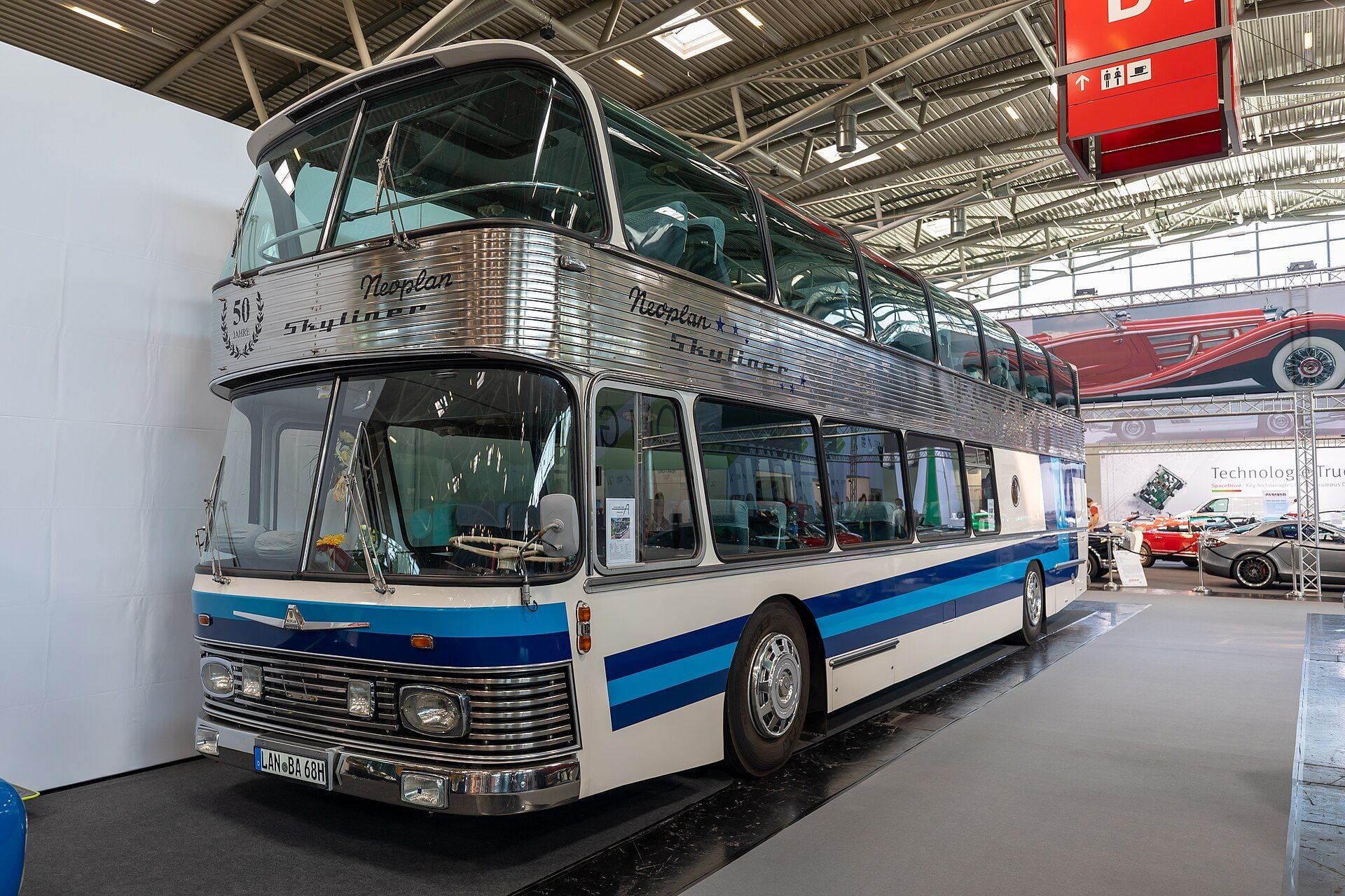 Older white and blue Neoplan Skyliner double-decker with a ribbed stainless-steel band, Neoplan Skyliner script and a 50 Jahre emblem, shown indoors at IAA Mobility 2021 in Munich