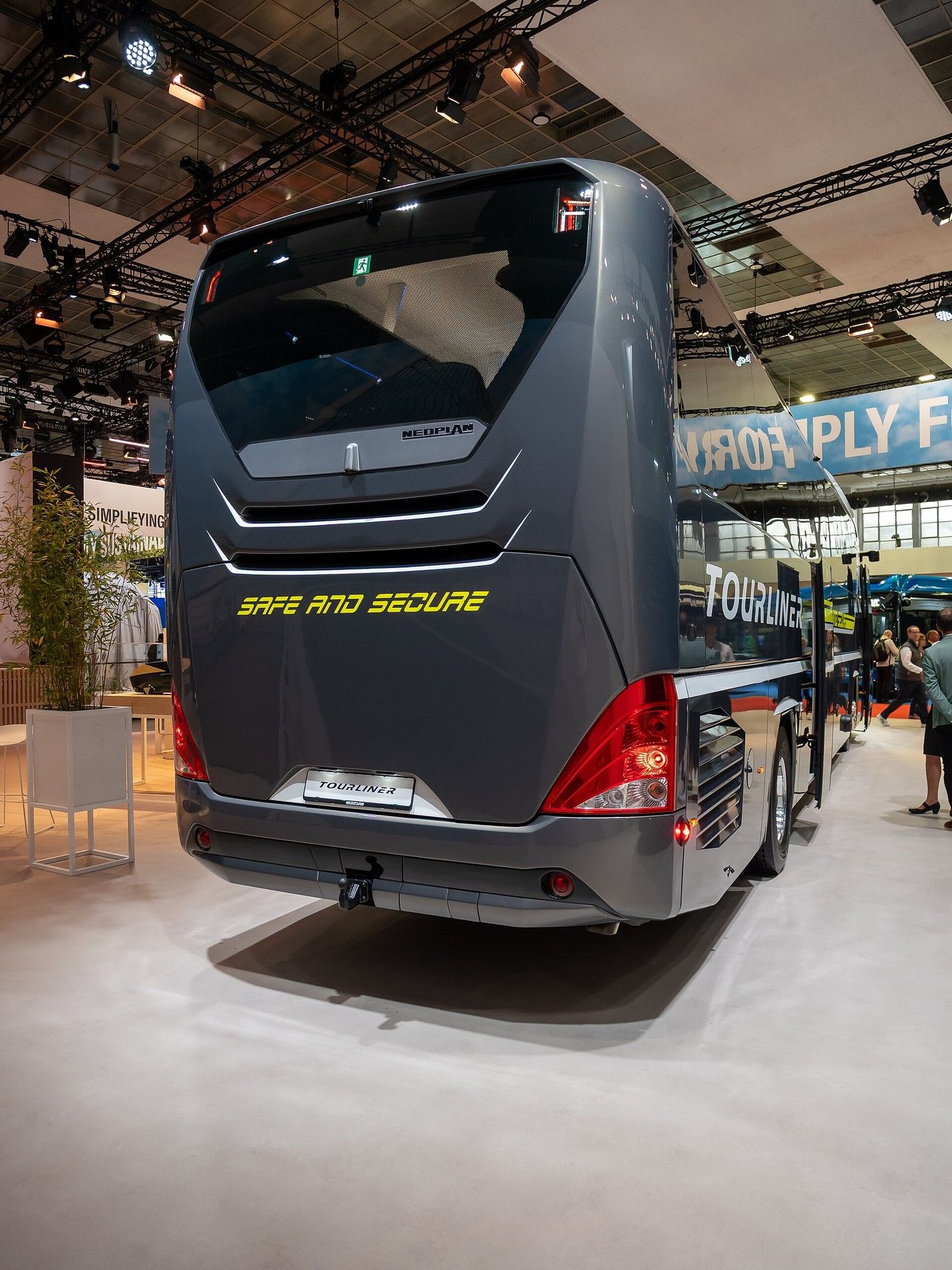 Rear of a dark grey Neoplan Tourliner with Safe and Secure lettering and a TOURLINER plate, shown indoors at Busworld Europe 2025 in Brussels
