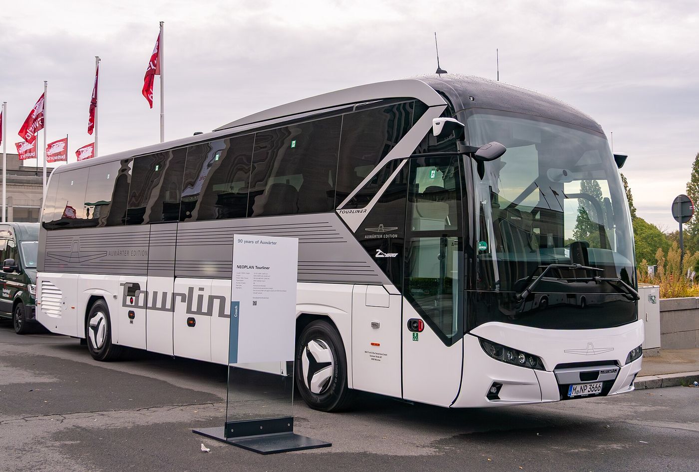 Silver and white Neoplan Tourliner two-axle touring coach with Auwärter Edition lettering, parked outdoors at Busworld Europe 2025 in Brussels with an information stand beside its front wheel